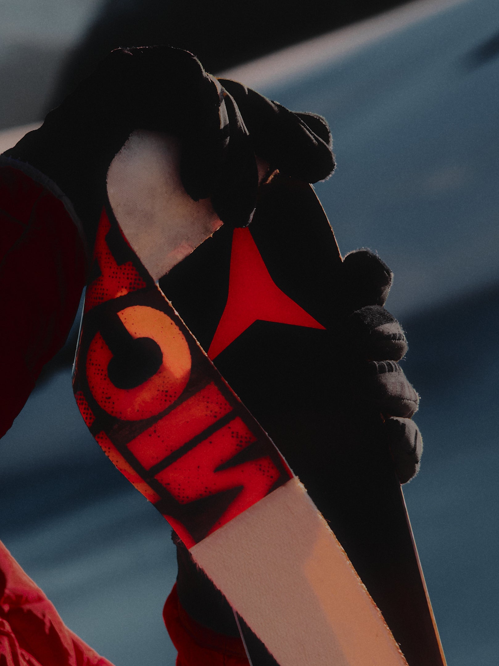 A close-up of a person wearing black gloves holding a black ski or snowboard with bold red and orange graphics, including a large stylized 'G' and a red star, against a blurred blue-gray outdoor background.