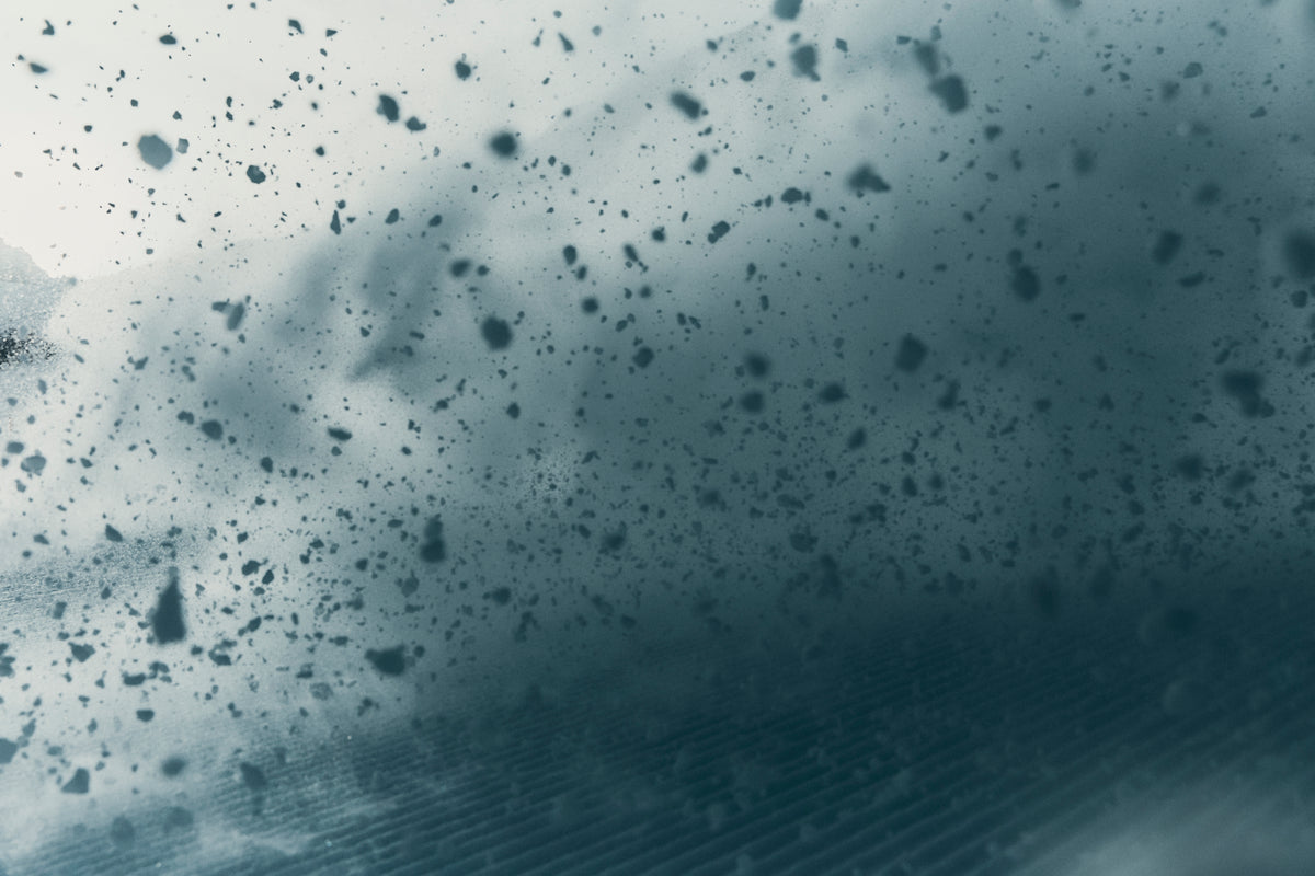 A close-up of a dark blue-gray textured surface covered with scattered small black specks or flecks, with a slightly blurred streaked pattern along the bottom edge.