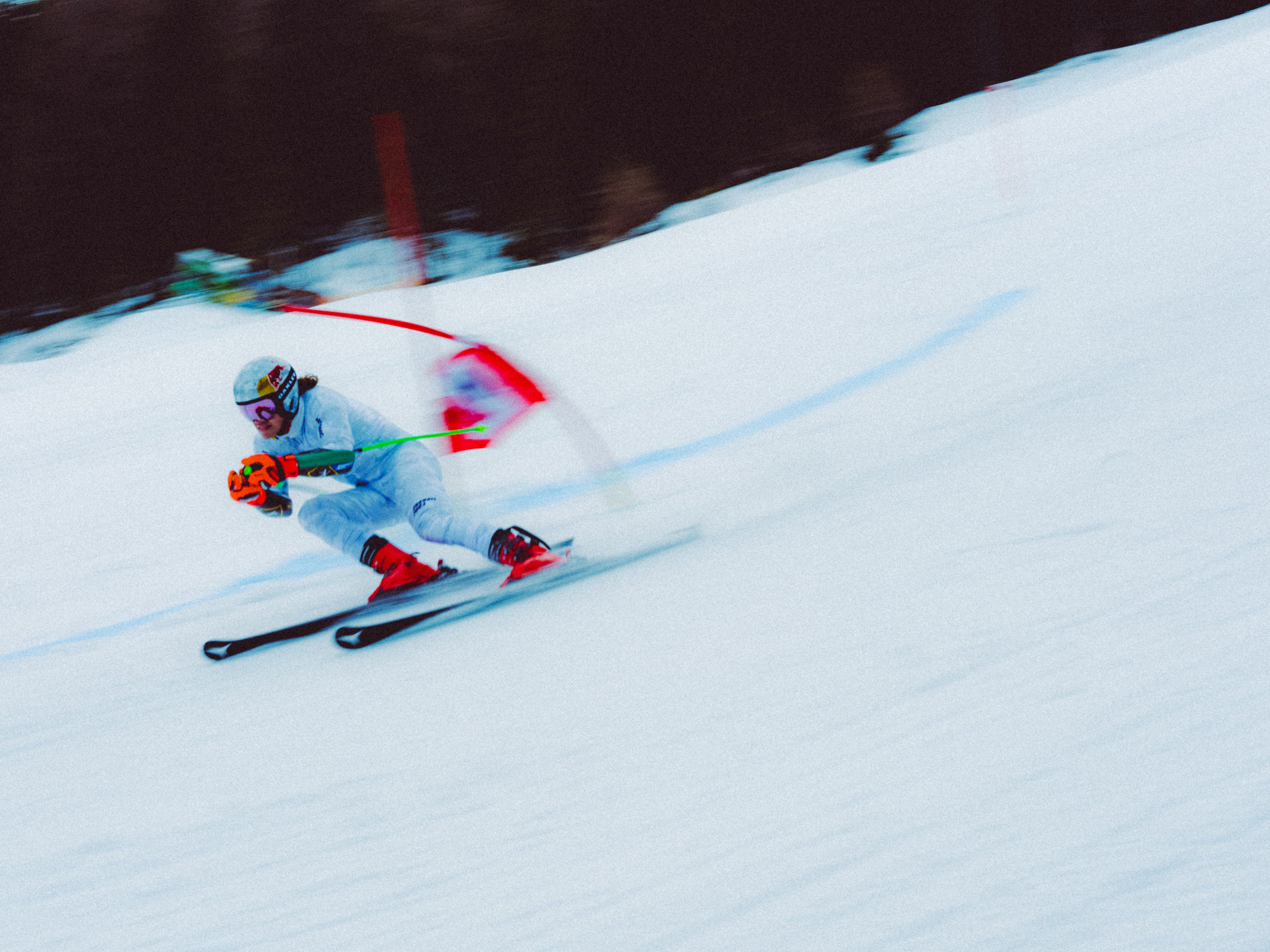 Lucas Pinheiro Braathen in a light gray suit and red boots carving through deep snow, holding ski poles with red, green, and orange accents. The background shows a blurred dark treeline and the white snow slope.
