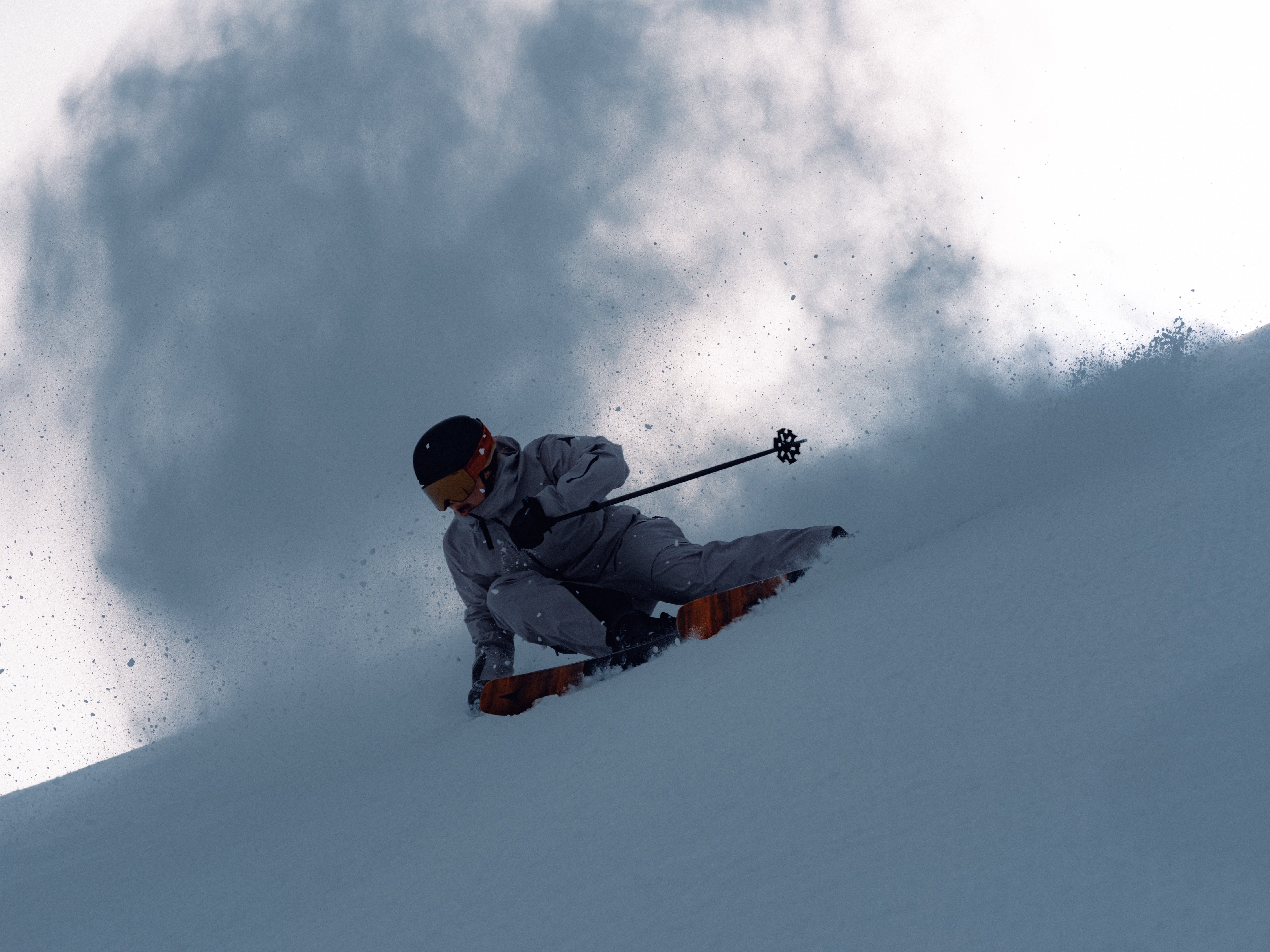 A skier in a light gray ski suit and black helmet carving through deep powder snow, kicking up a large spray of snow behind him.
