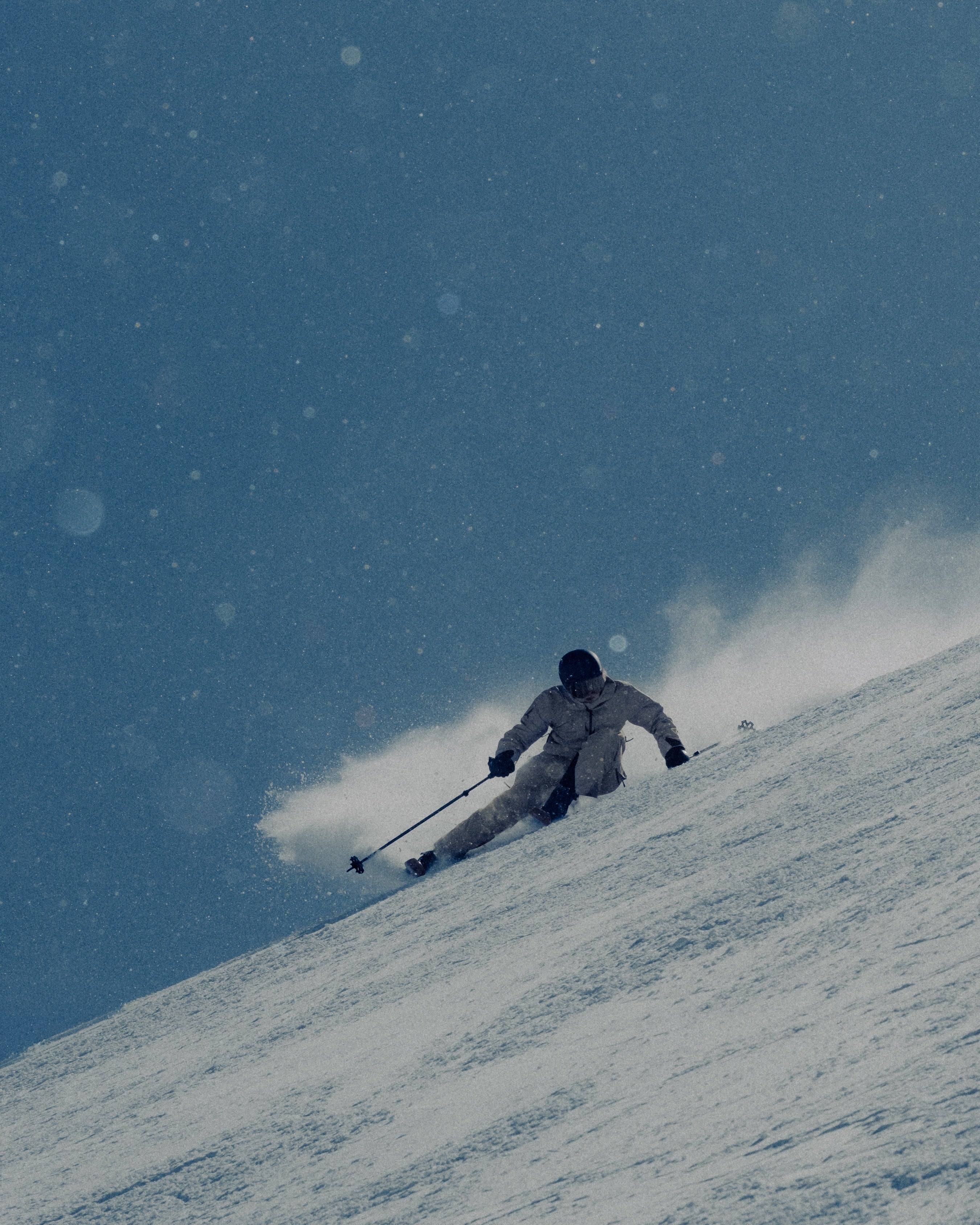 A skier in a light-colored suit carving through fresh powder on a snowy slope, with a spray of snow kicked up behind them under a blue sky.