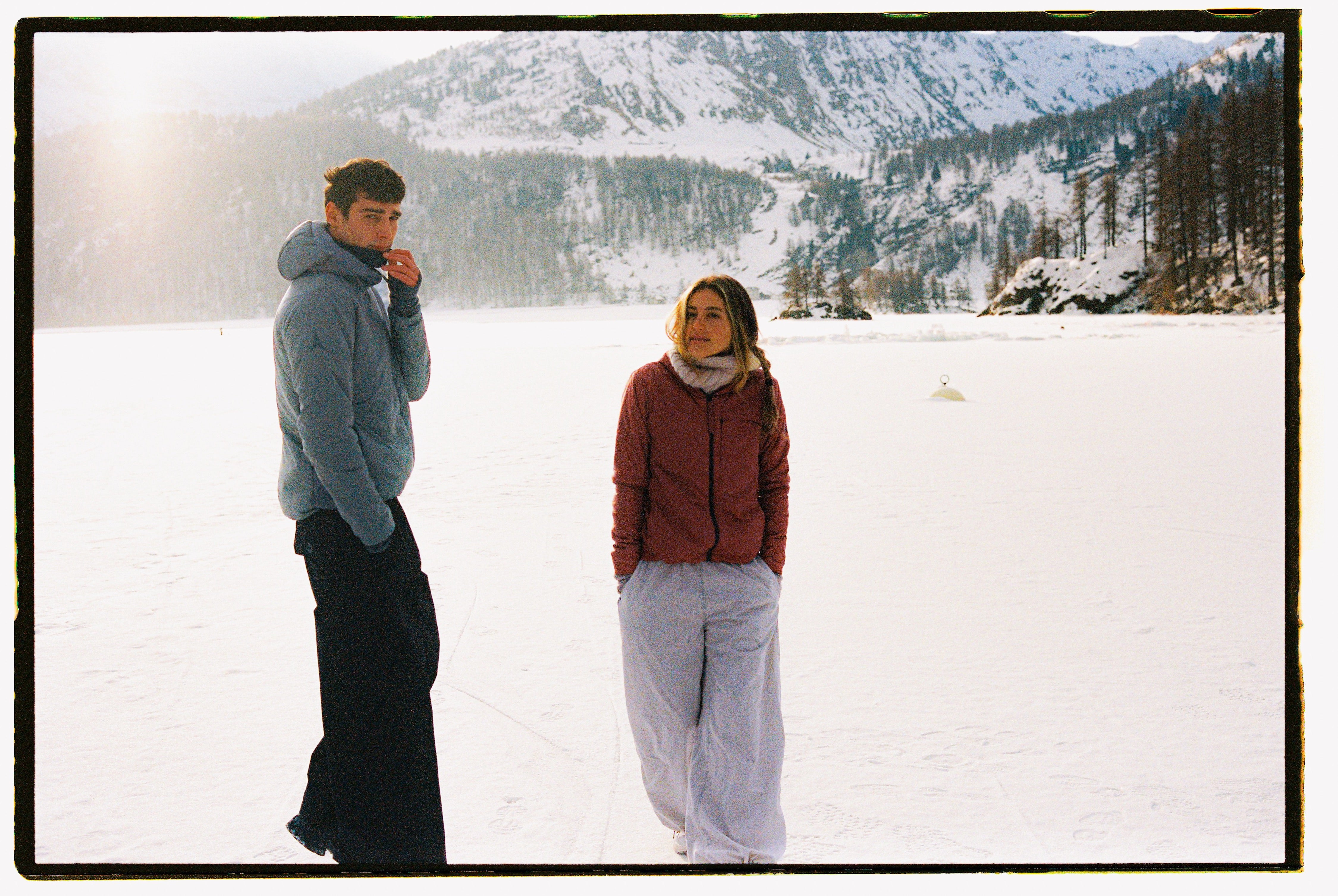 A man and a woman standing on a snowy mountain landscape. The man wears a light gray puffer jacket and black pants, while the woman wears a rust-red puffer jacket and light gray pants.