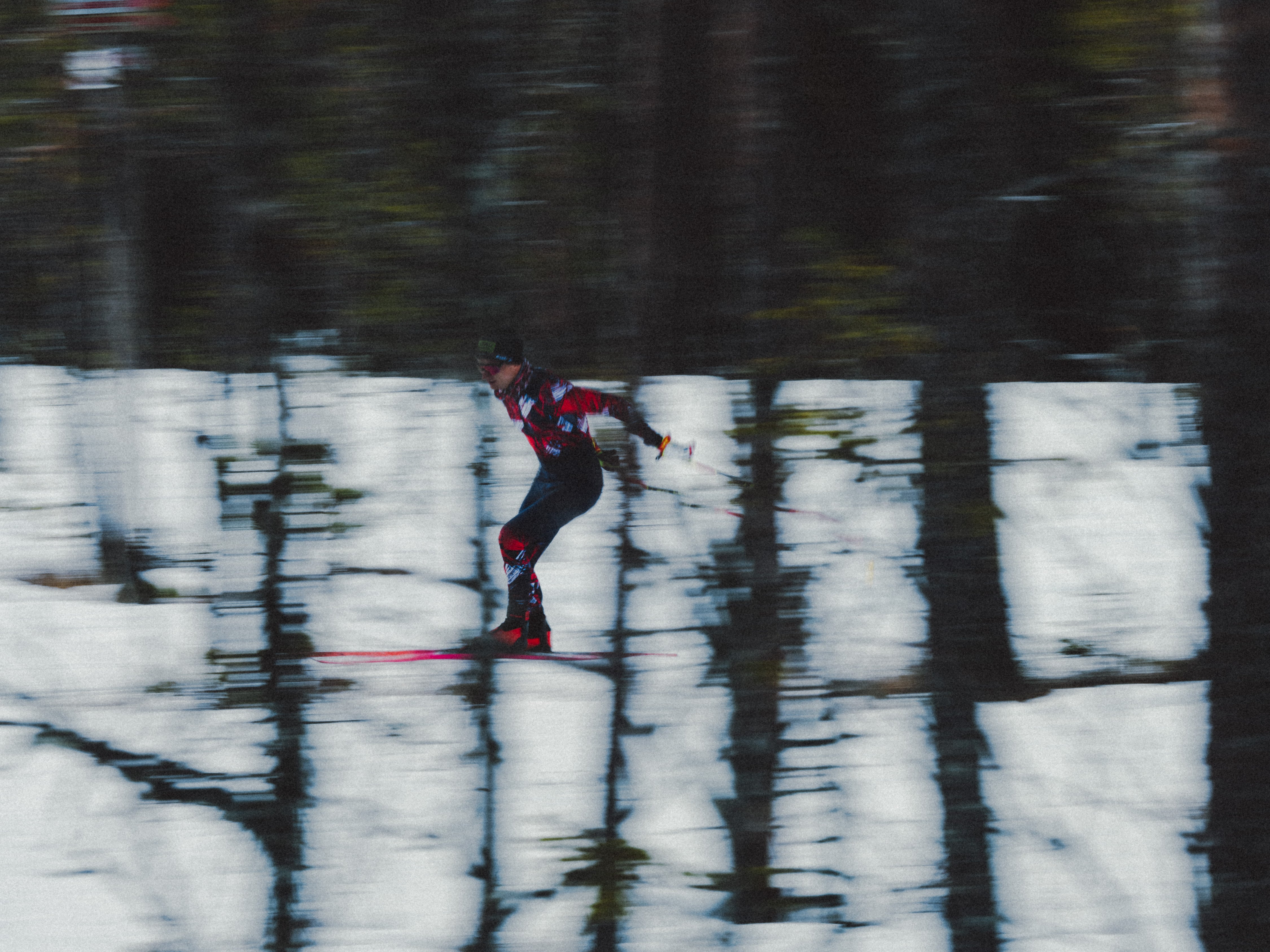 Mika Vermeulen gliding across a snowy surface with blurred tree reflections in the background, wearing a red and black ski suit.