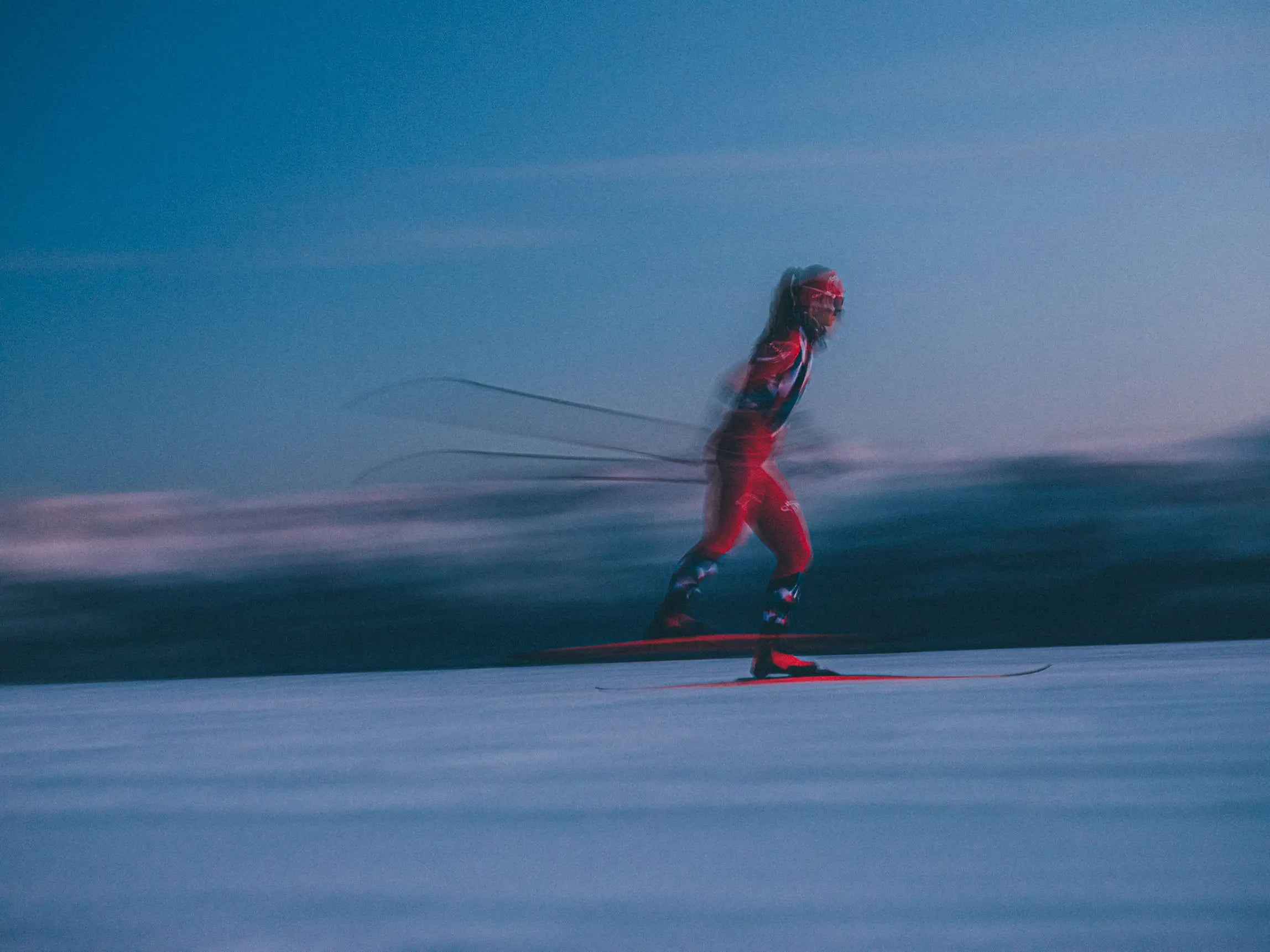 A skier in a red and blue racing suit glides across a snowy slope under a clear blue twilight sky, captured with motion blur around the body and ski poles.