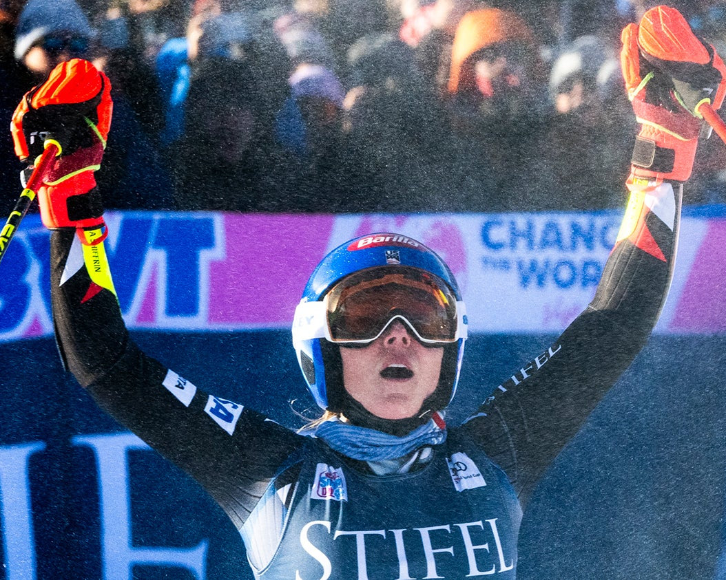 Mikaela Shiffrin celebrates with arms raised after a race, wearing a blue helmet, goggles, and a black racing suit with 'STIFEL' on the chest, holding ski poles in a snowy outdoor setting.