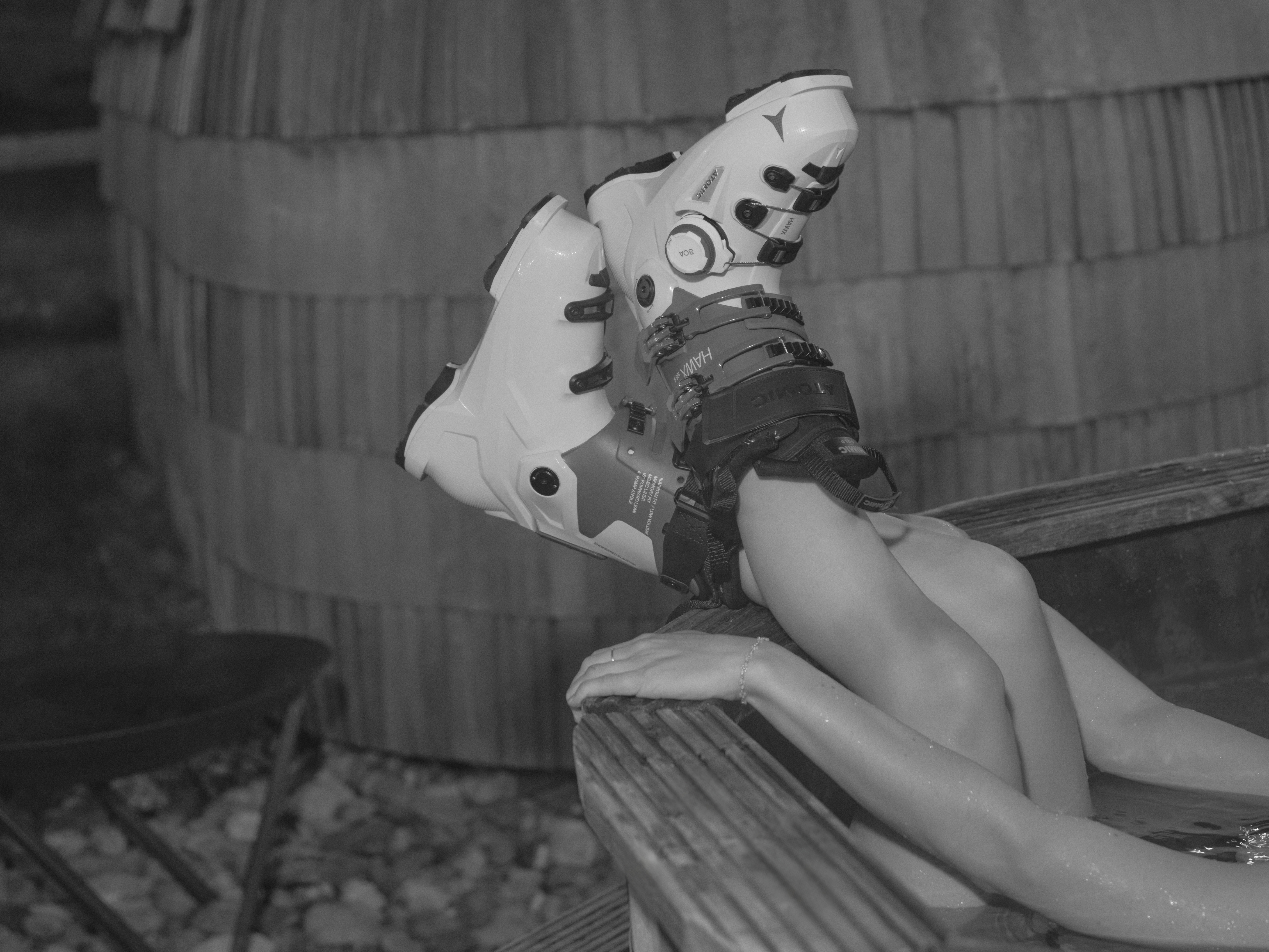 Black-and-white photo of a person relaxing in a wooden outdoor hot tub with legs raised and wearing white Atomic Hawx ski boots. Large wooden barrels and stones in the background create an alpine spa atmosphere.