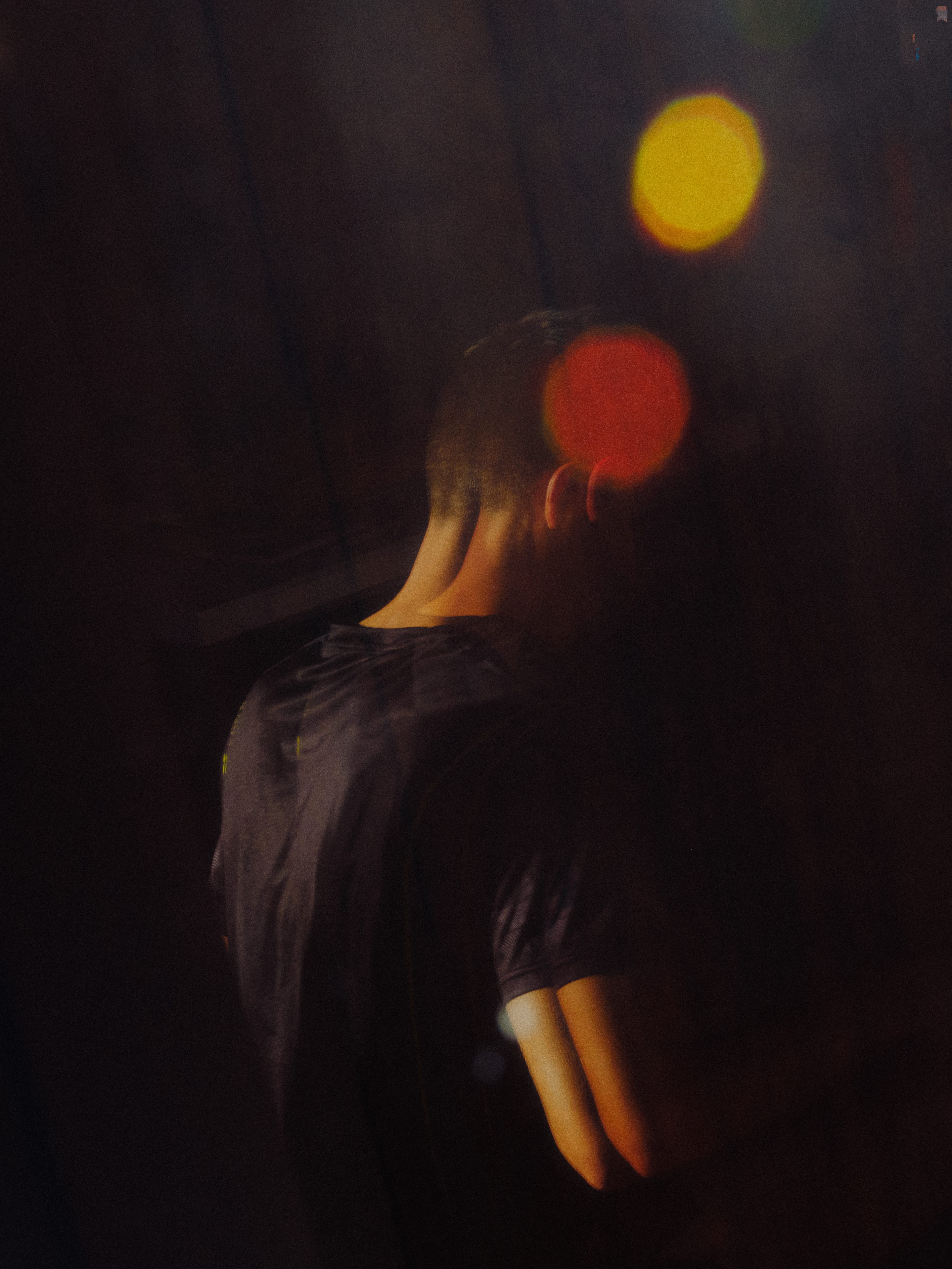 A person seen from behind wearing a dark t-shirt, standing in a dimly lit environment with warm yellow and orange bokeh light effects.