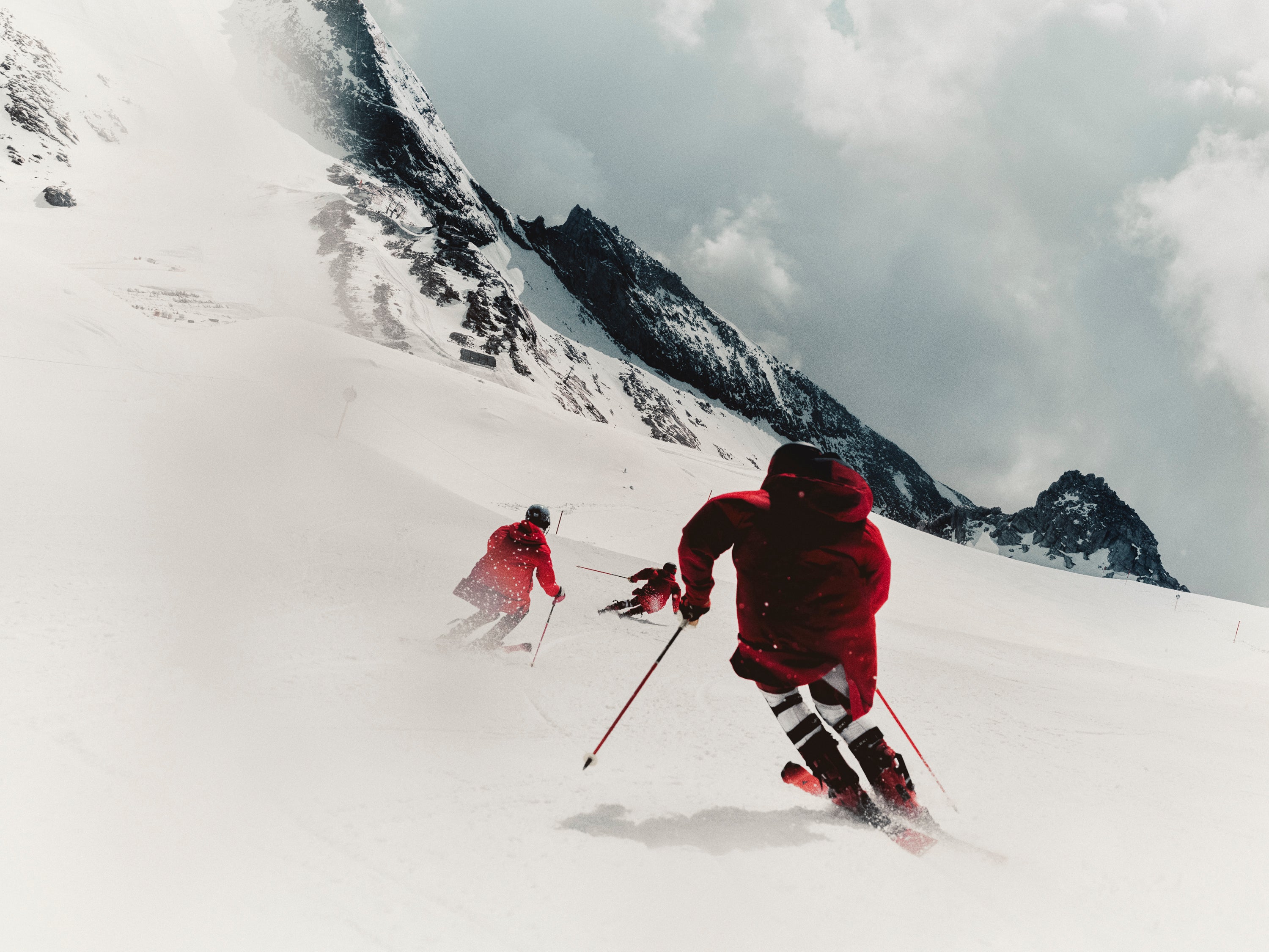 Two skiers descend a snowy mountain slope, with one skier in the foreground wearing a bright red jacket and black pants, and another skier further behind in a matching red jacket. Jagged dark mountains and a cloudy sky are visible in the background.