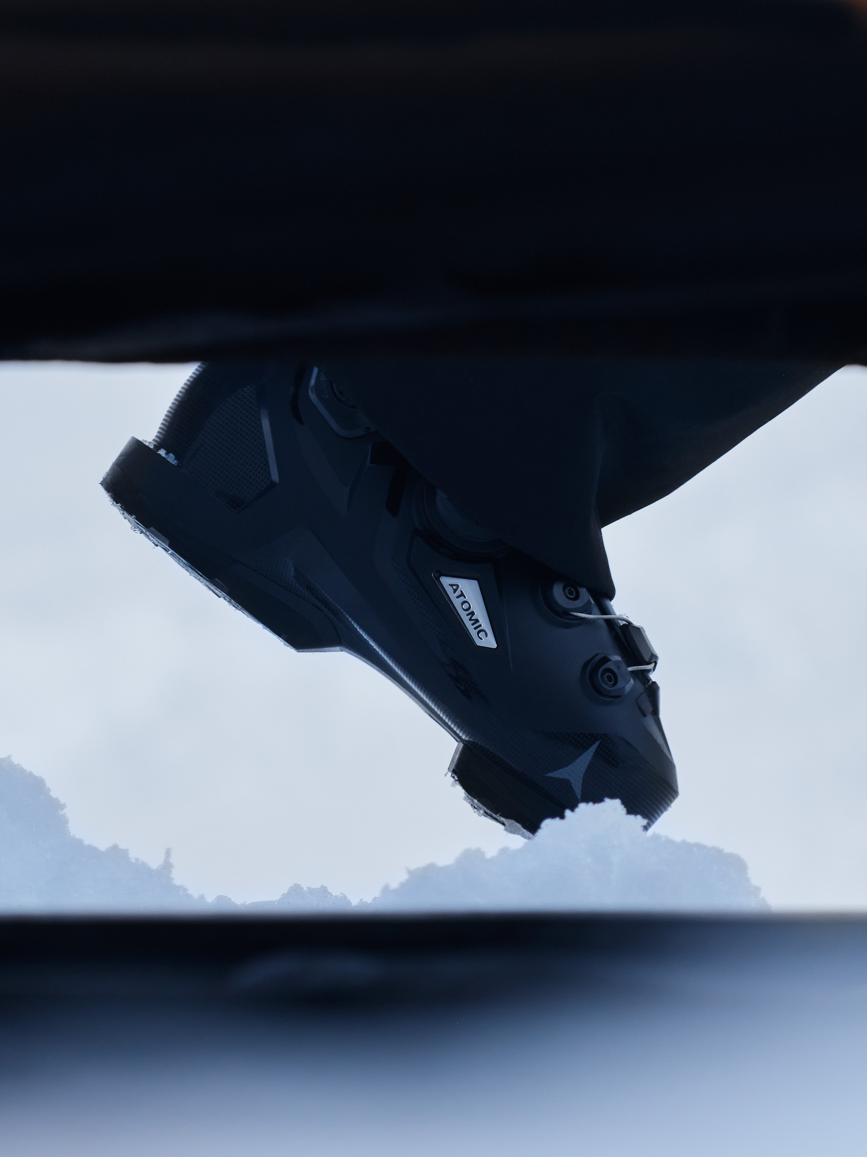 A close-up, low-angle shot of a black Salomon ski boot hanging upside down against a snowy mountain background.