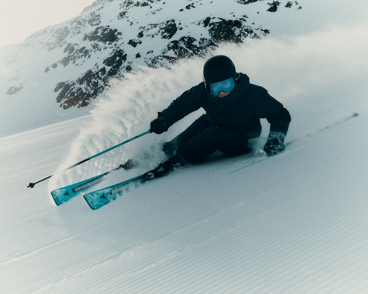 A skier in a black outfit and blue goggles carves through fresh powder snow on a mountain slope, with snow spraying around them and rocky mountains in the background.