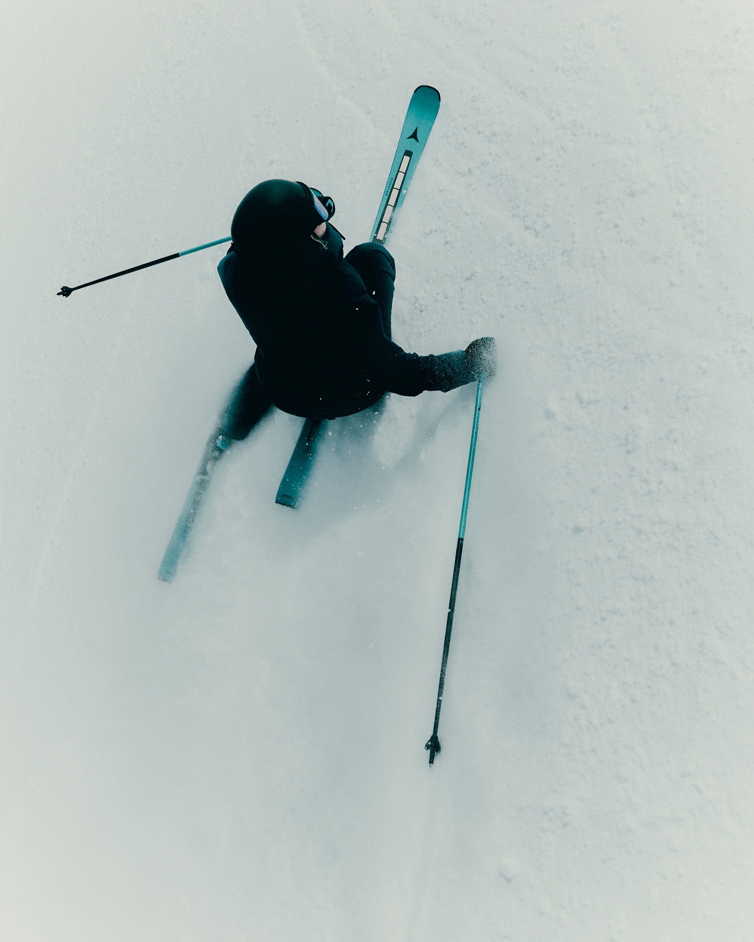 A skier in a black outfit carving through deep powder snow, viewed from above, with ski poles and skis visible against a bright white snowy background.