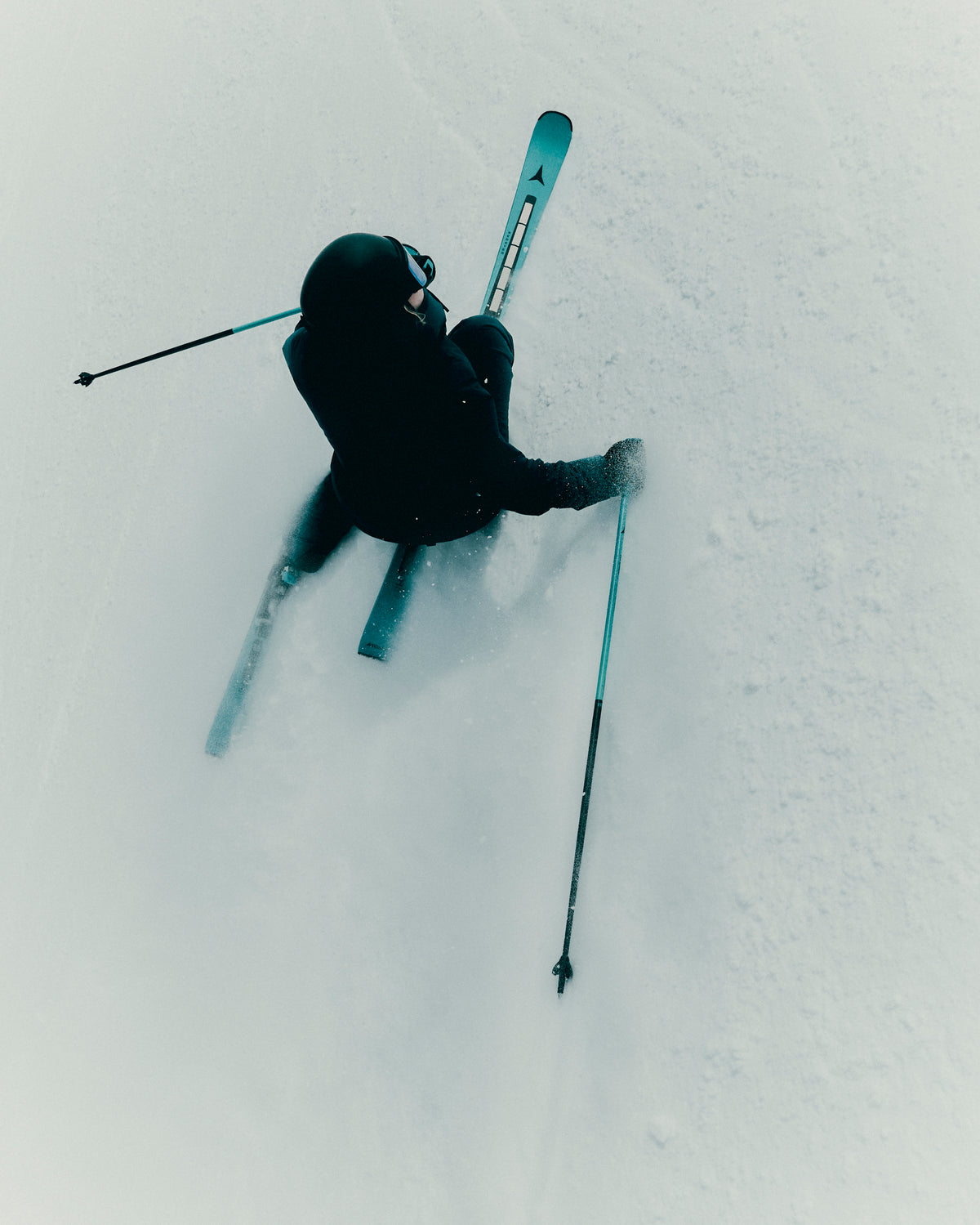 A skier in a black outfit carving through deep powder snow, viewed from above, with ski poles and skis visible against a bright white snowy background.