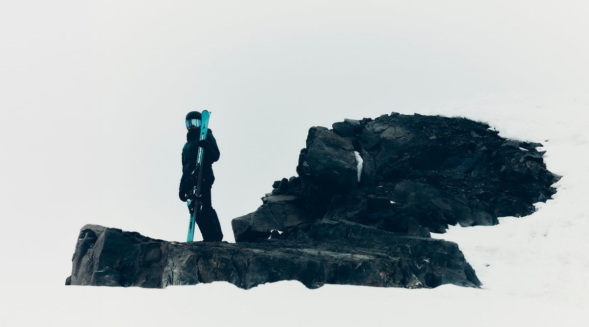 A person in dark winter clothing stands on a snowy rock holding bright turquoise-blue ski poles or skis against a pale sky, with dark rocks behind them.