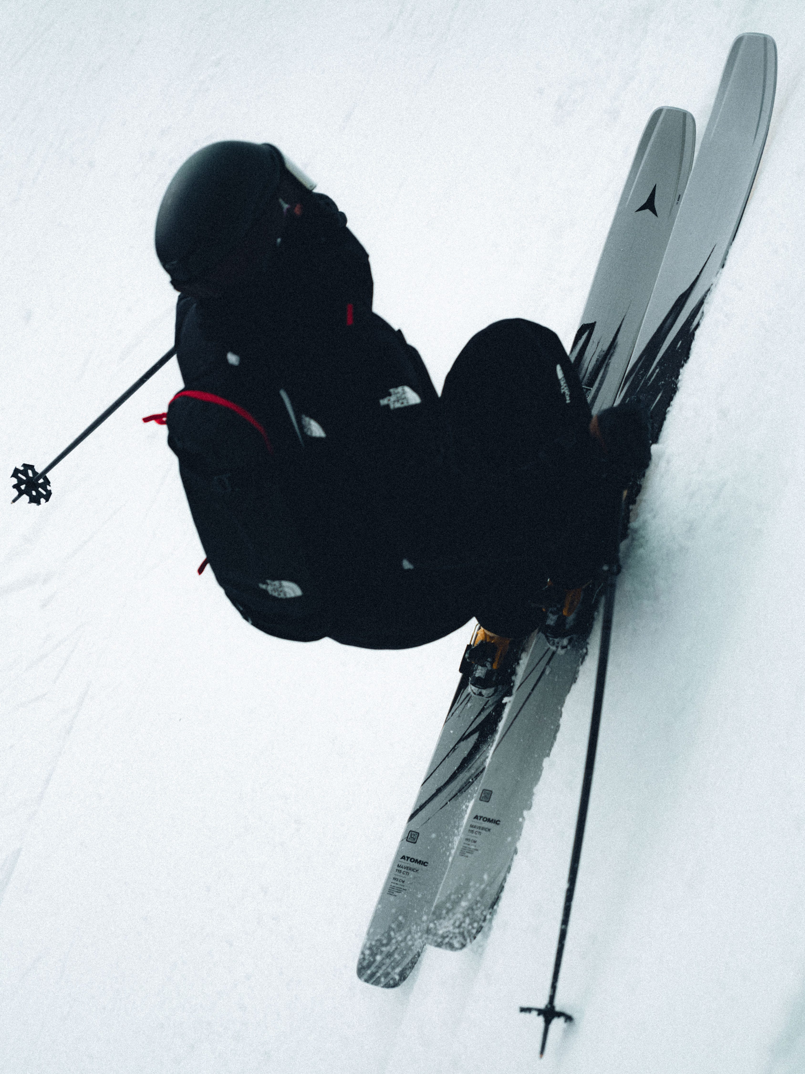 A skier in a black ski suit and helmet is captured mid-action on a snowy slope, with skis angled vertically and ski poles visible against a bright white snow background.