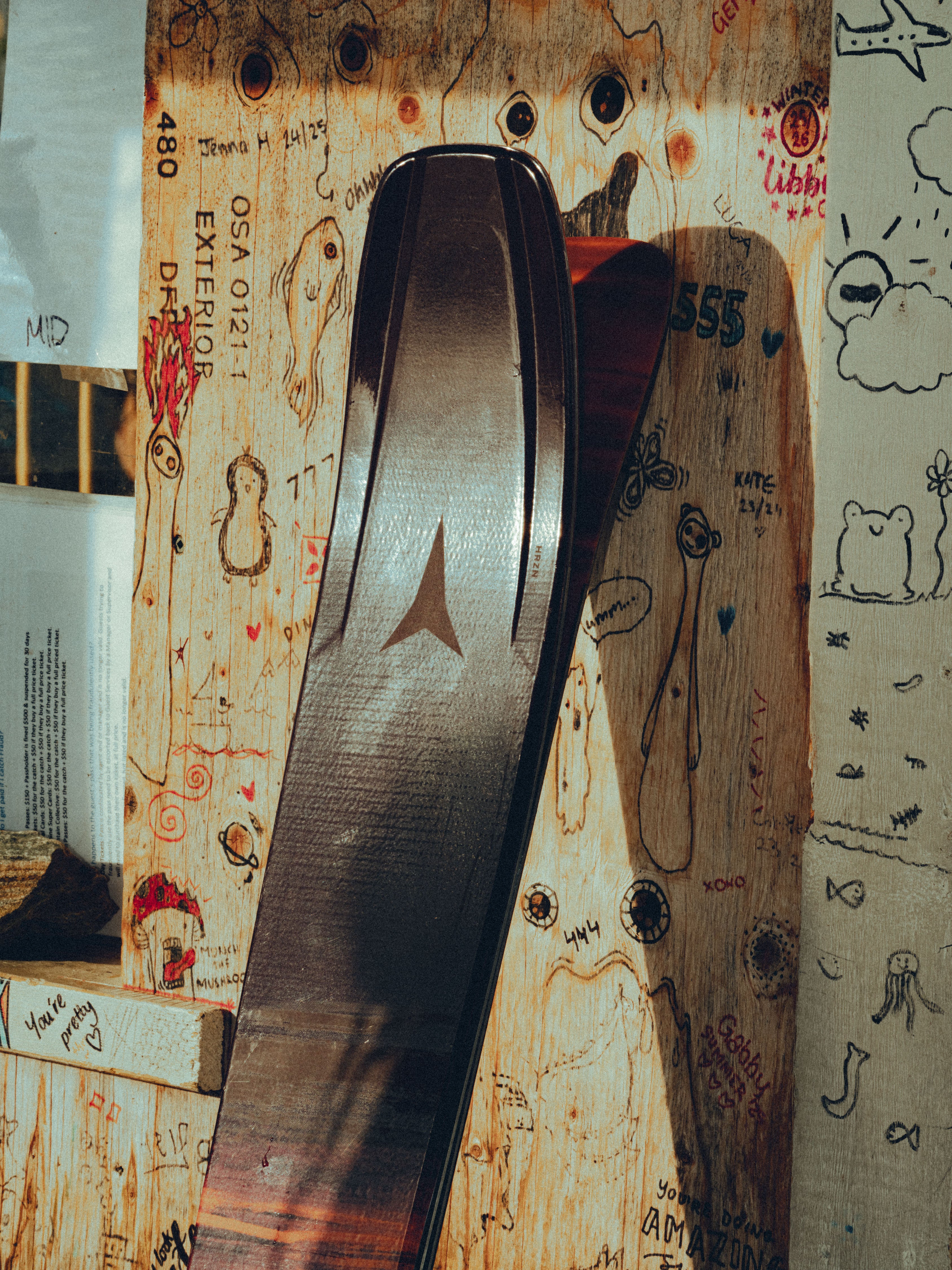 A close-up of a dark brown wooden ski or snowboard blank leaning against a graffiti-covered wooden wall.