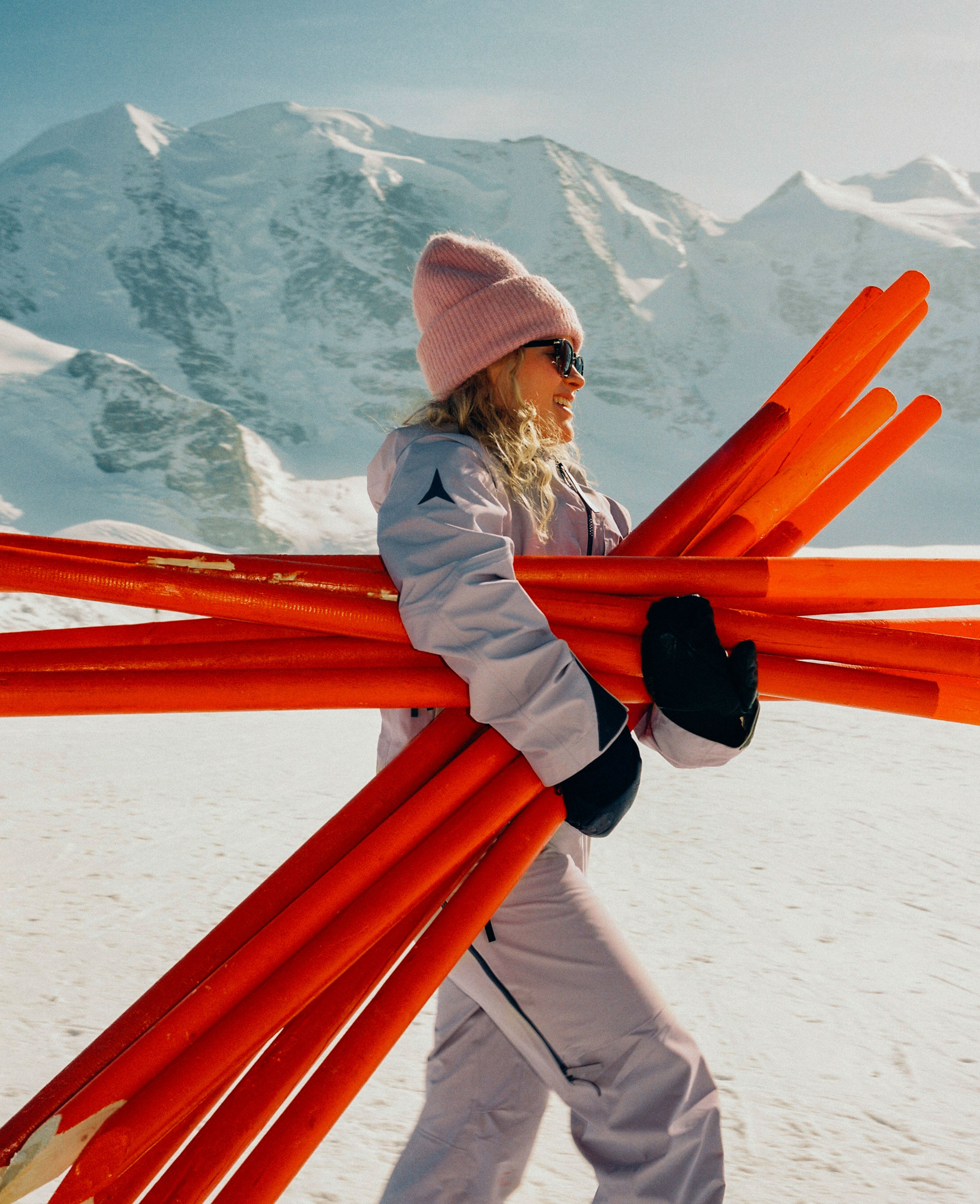 A woman in ski gear carrying a bundle of bright orange skis in a snowy mountain setting.