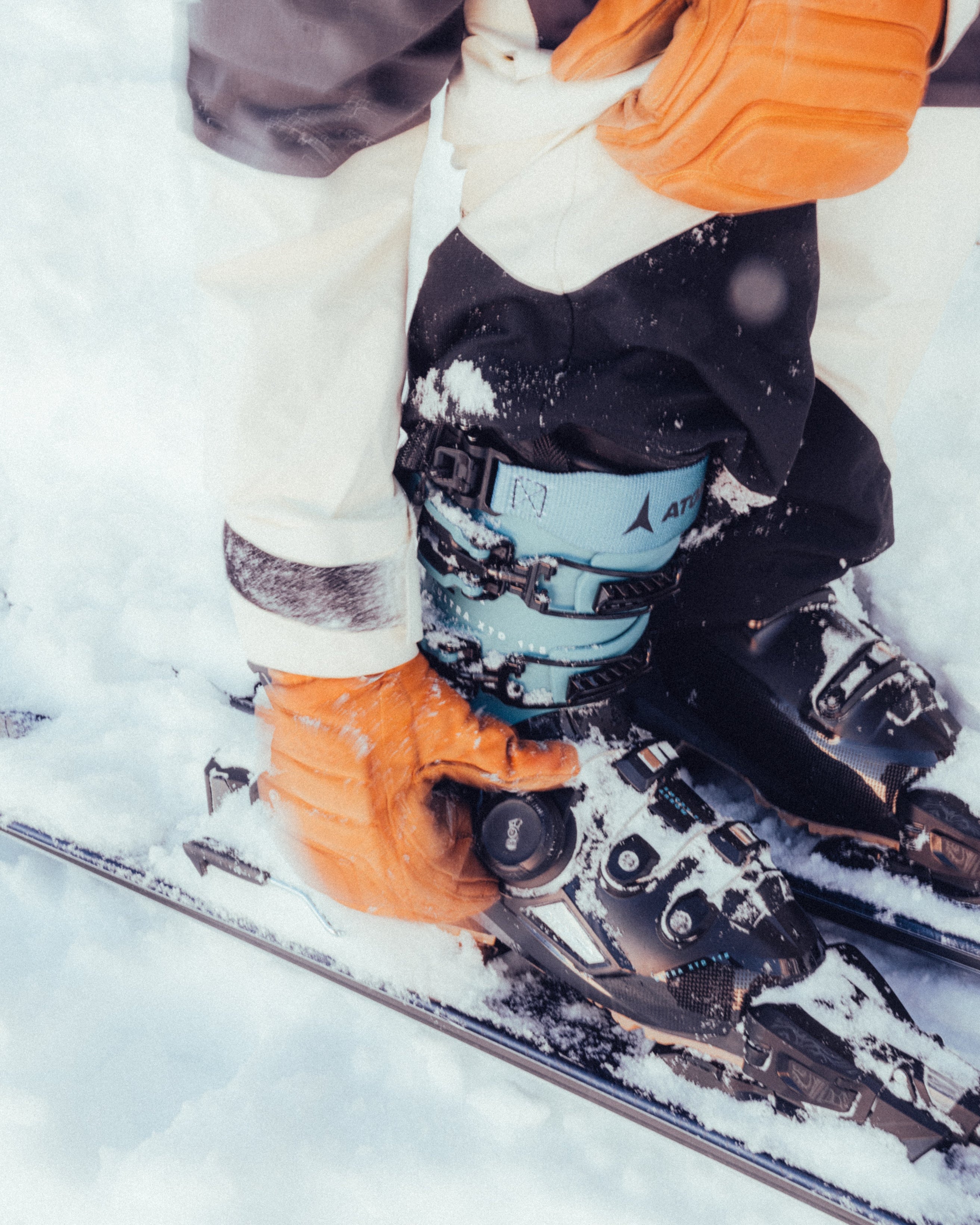Close-up of a skier adjusting bindings on a ski boot in the snow, wearing orange gloves and black ski pants.