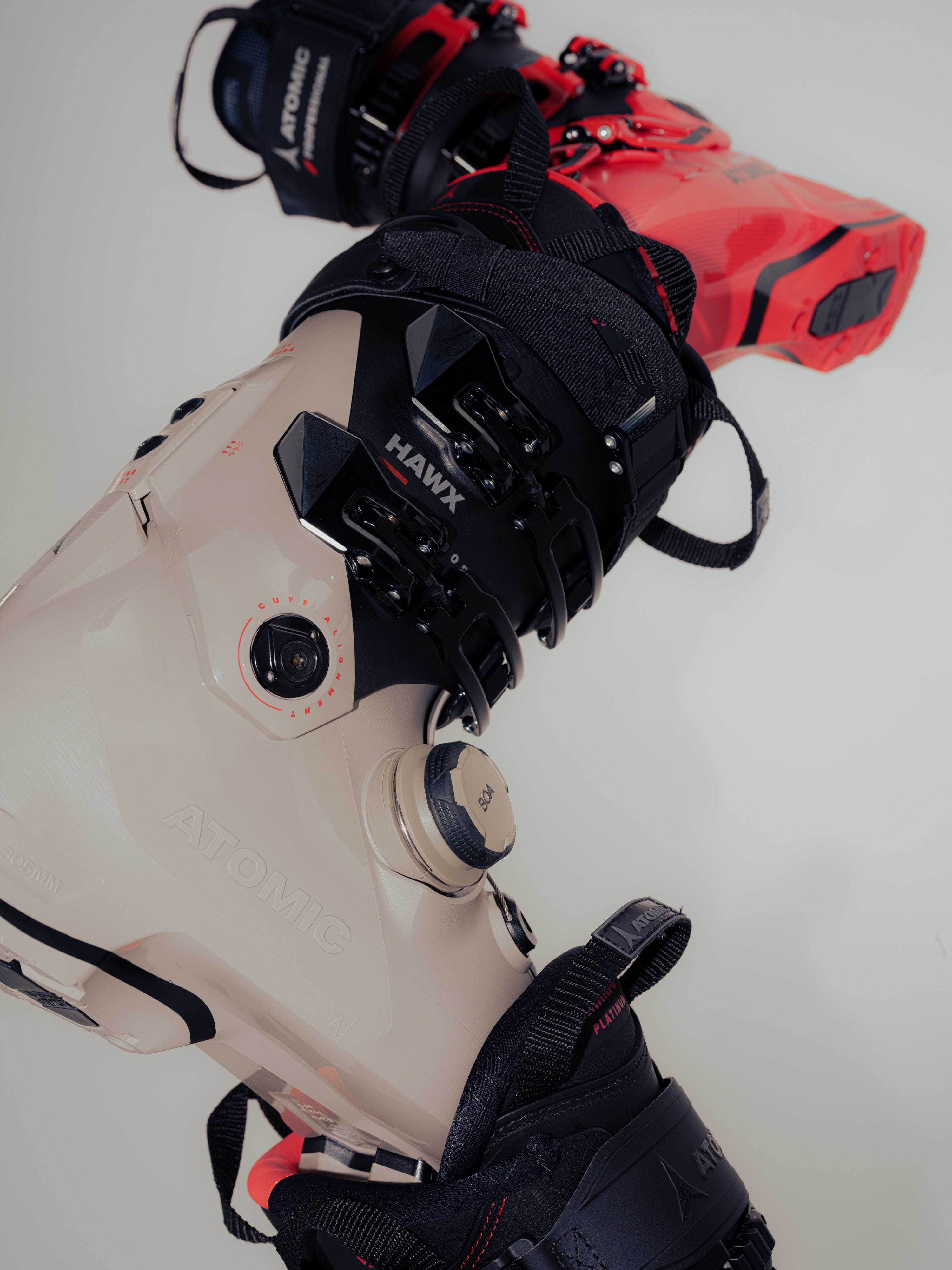Close-up of a ski boot with a beige Atomic shell, black Hawk buckle system, and red and black ski bindings attached, photographed against a plain light background.