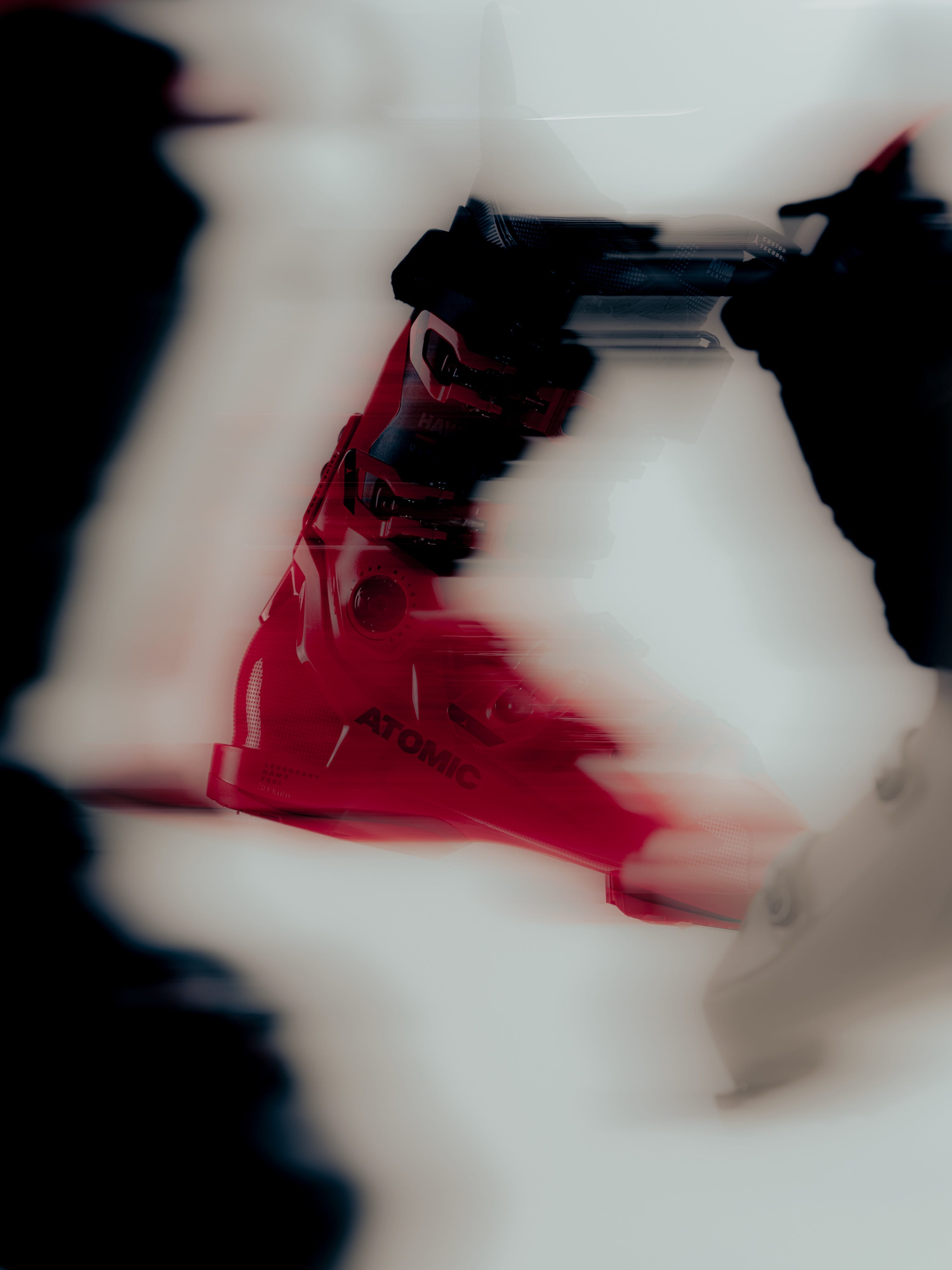A red ski boot with black buckles and straps, shown in a heavily blurred, stylized close-up with dark silhouettes framing the scene.