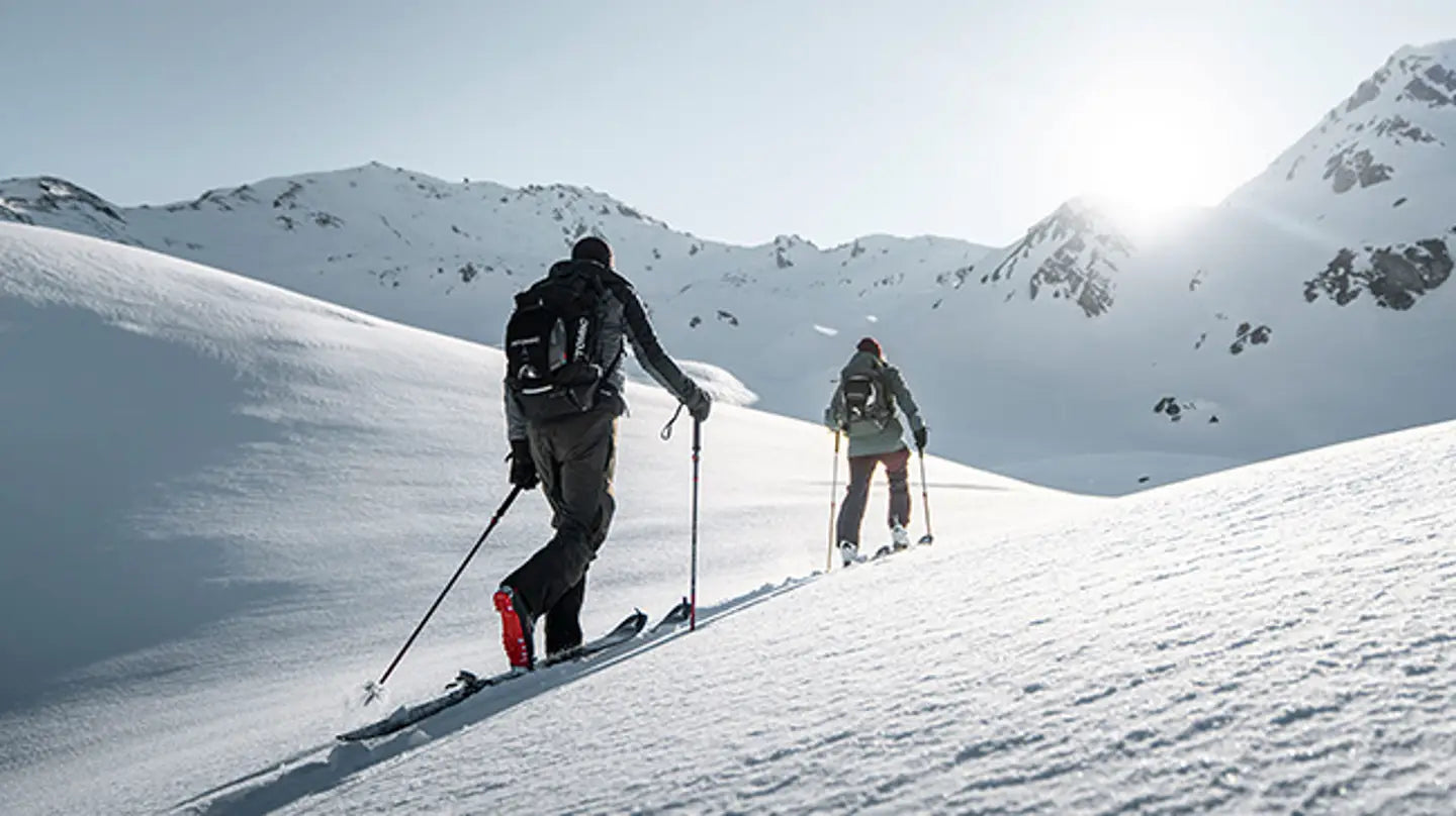 Two skiers traverse a snowy mountain slope under a bright sun, with snow-covered peaks in the background.