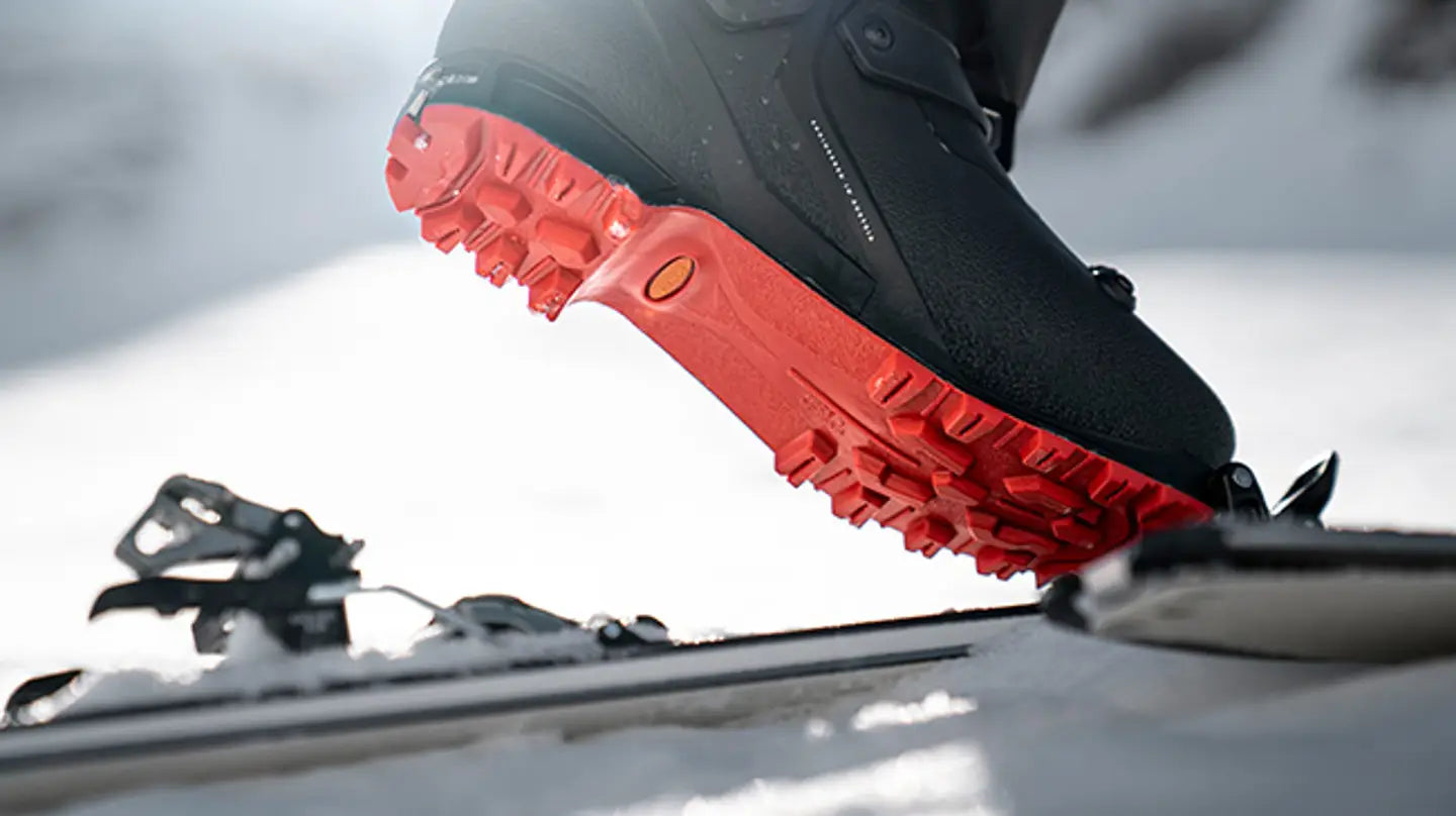 A close-up of a black ski boot with a bright red sole clipped onto a touring binding in a snowy outdoor setting.