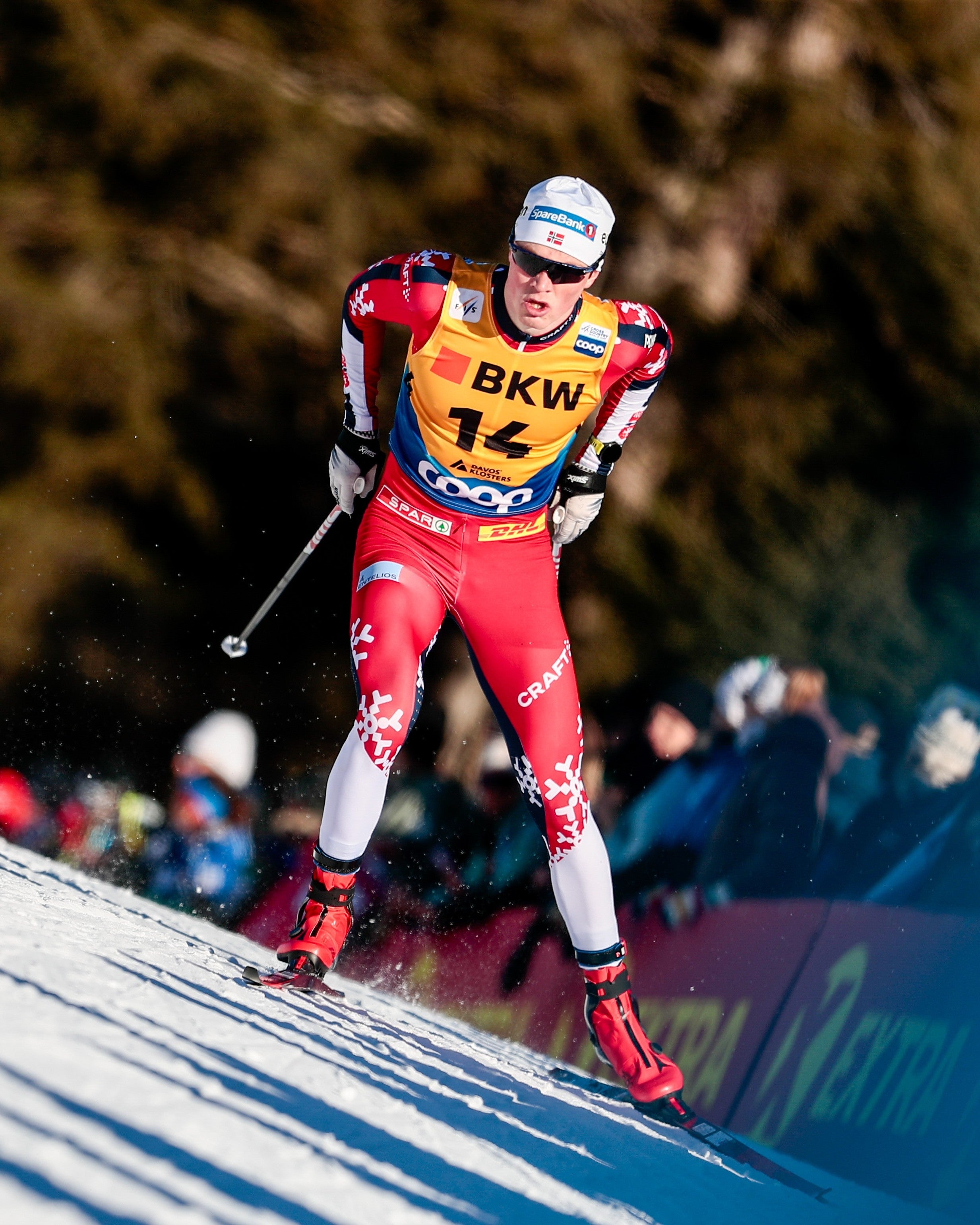 A cross-country skier in a red, white, and yellow racing suit with the number 14 and 'BKW' on the chest, skiing down a snowy slope with trees and spectators blurred in the background.