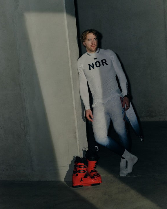 Aleksander Aamodt Kilde wearing a white race suit with 'NOR' printed on the chest stands in a dim concrete room, with red ski boots resting on the floor nearby.