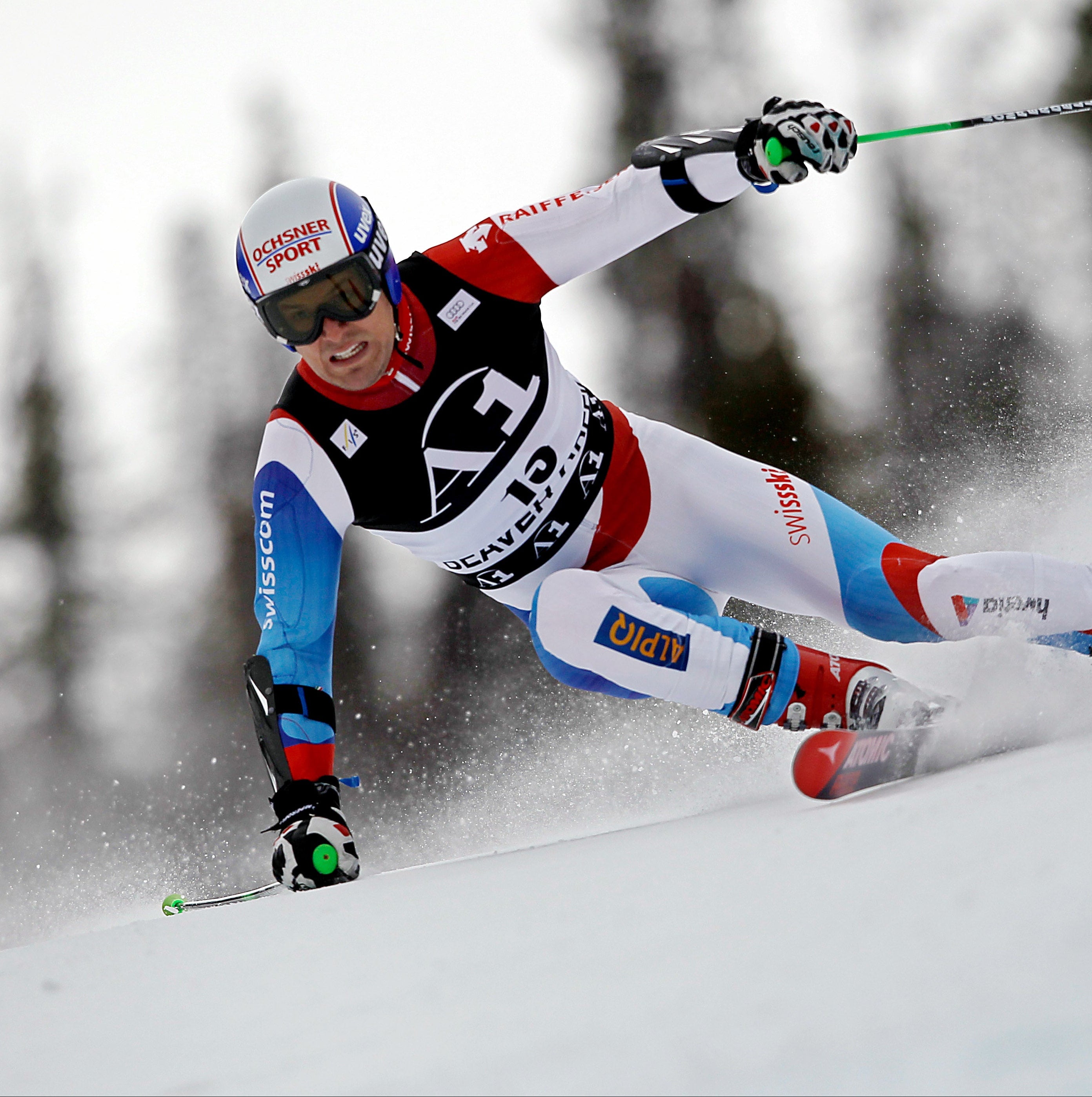 Daniel Albrecht in a red, white, and blue racing suit leans sharply into a turn on a snowy race course. The skier is wearing a white helmet with red and blue graphics, black goggles, and ski poles.