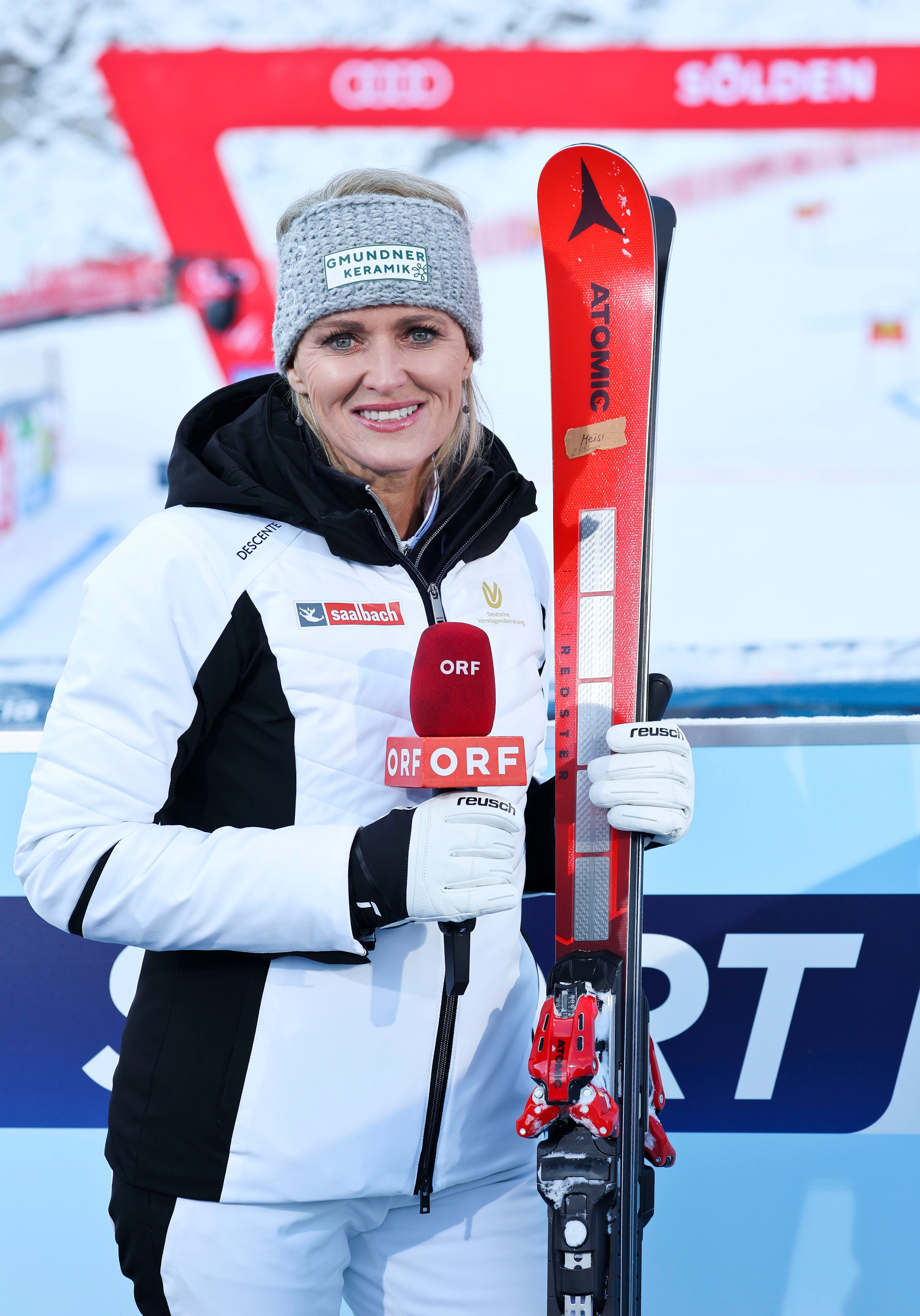 Alexandra Meissnitzer in ski gear stands outdoors holding red skis and an ORF microphone. She wears a white and black ski jacket, white ski pants, a gray knit beanie, and white gloves.