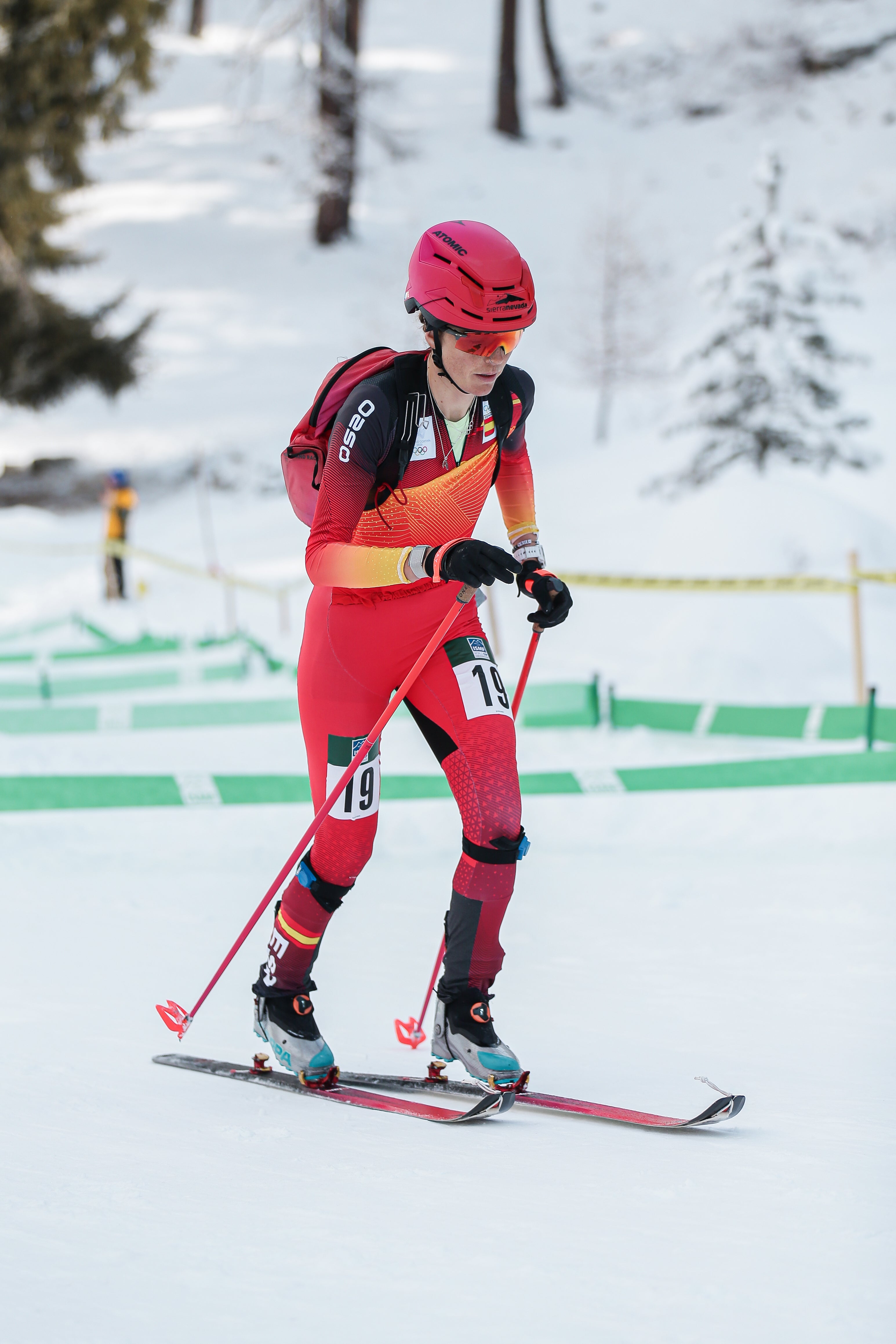 Ana Alonso in a red and black racing suit with a red helmet and goggles, holding ski poles on a snowy track.