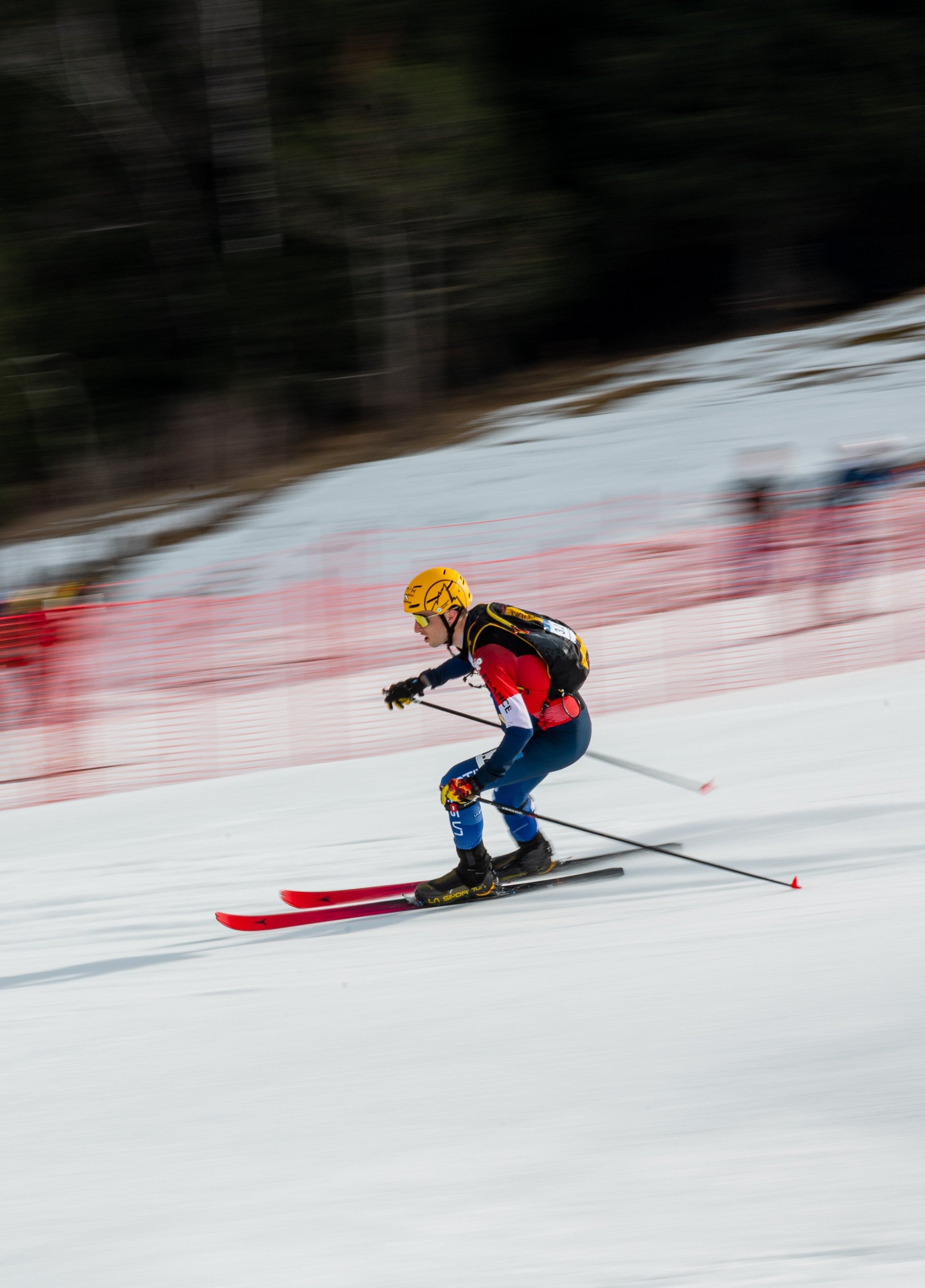 Thibault Aselmet in a red, black, and blue racing suit with a yellow helmet glides across a snowy slope with red safety fencing and blurred trees in the background.