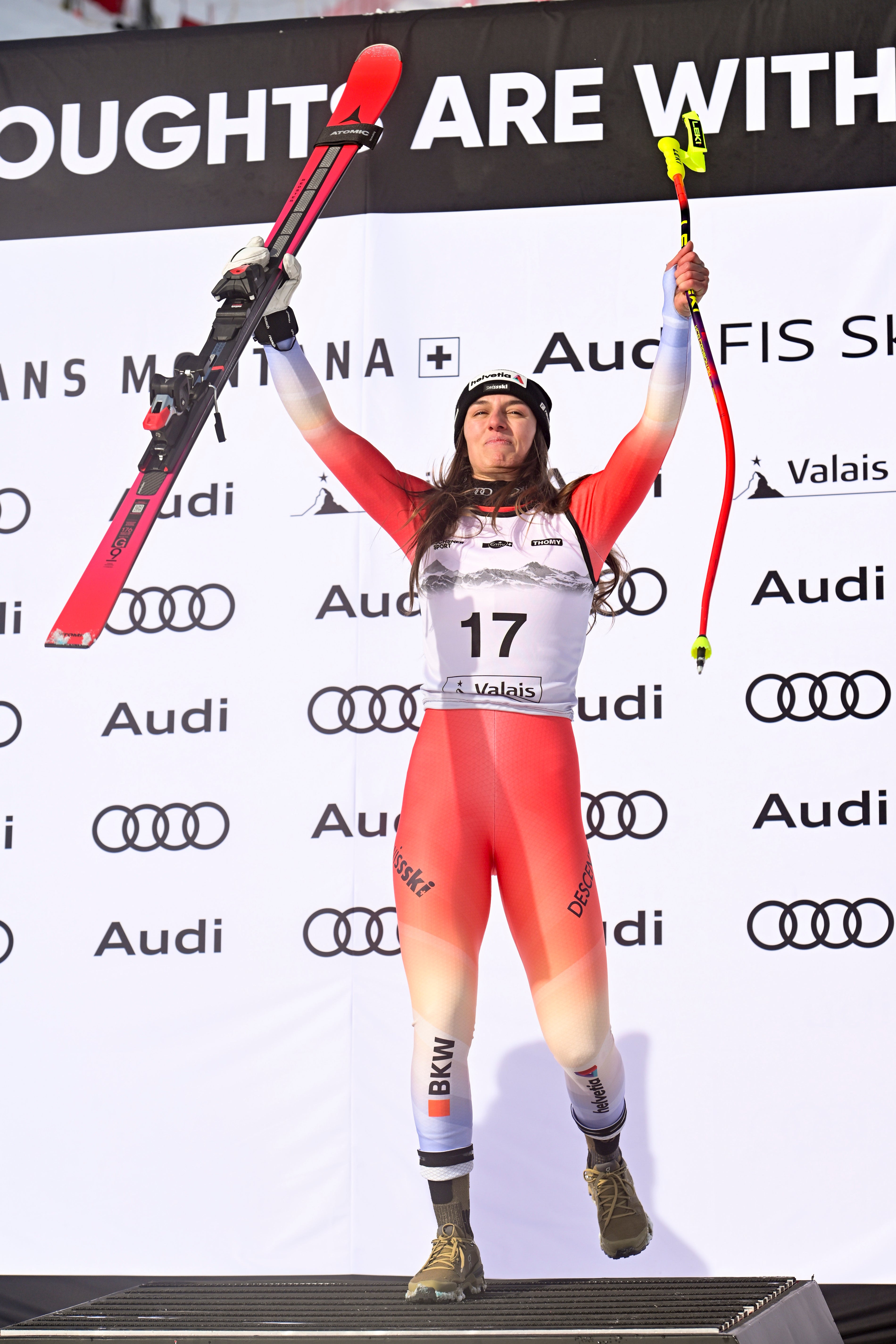 Malorie Blanc in a red, white, and gray ski suit jumps joyfully on a podium while holding up red skis and ski poles. The background features a white banner with repeated logos and text.