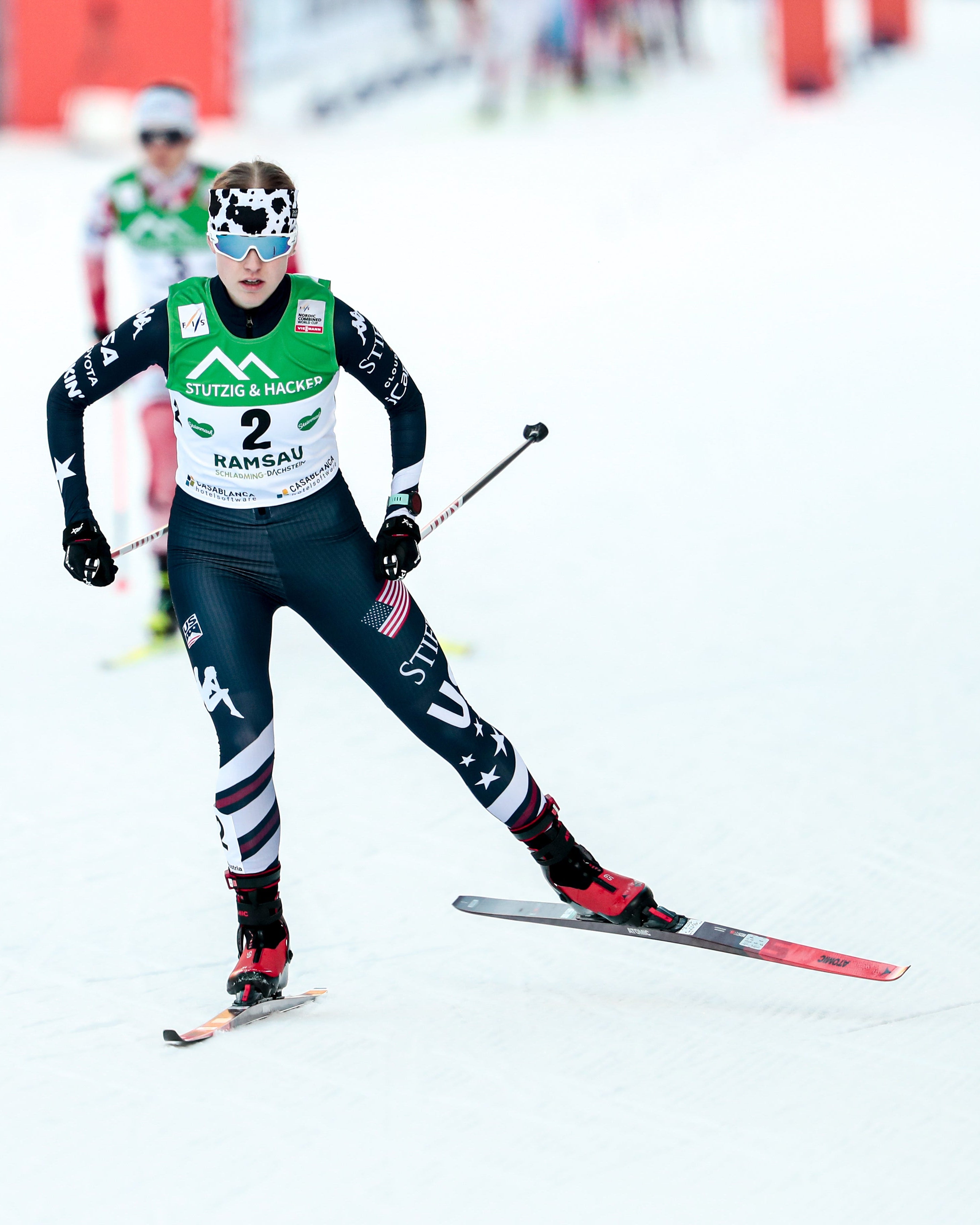 The cross-country skier Alexa Brabrec in a dark navy and green racing suit with bib number 2, skiing on a snowy track with poles and skis visible.