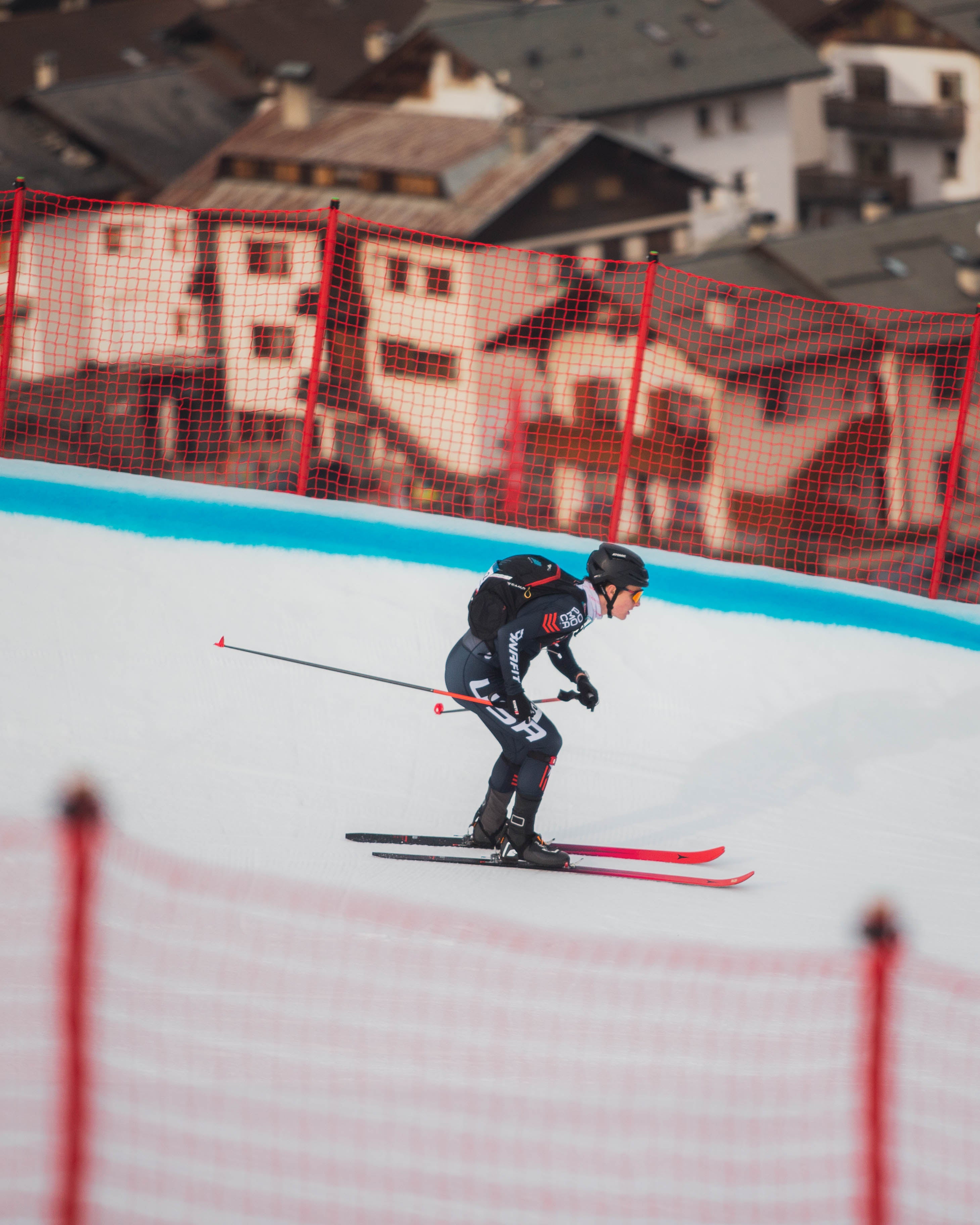Griffin Briley in dark clothing glides down a snowy slope with red skis and poles, with a red safety net and buildings in the background.