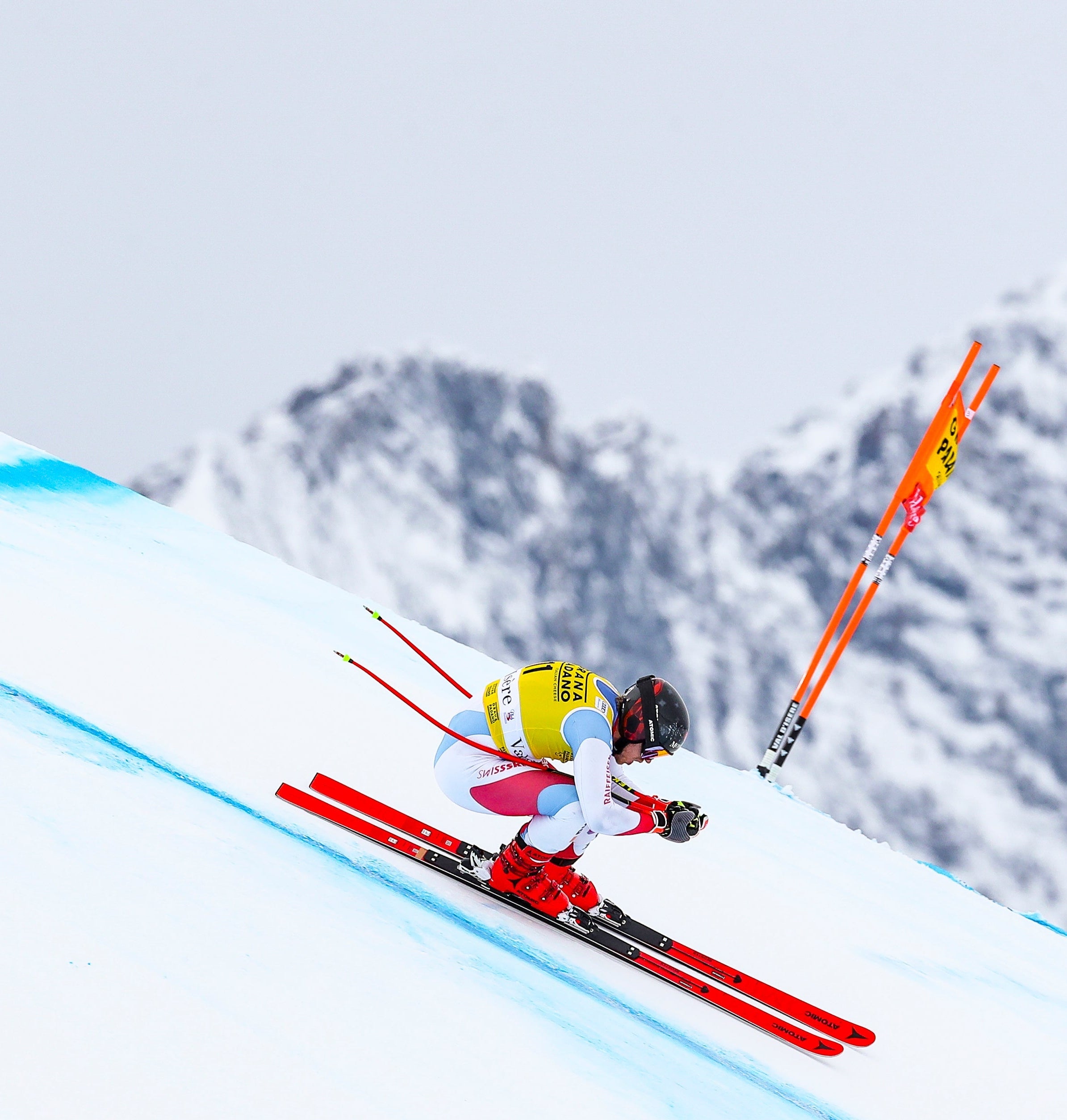 Mauro Caviezel in a bright yellow, white, and red racing suit leans sharply into a turn on a snowy mountain slope, with red skis and orange ski poles visible against a backdrop of snow-covered peaks.