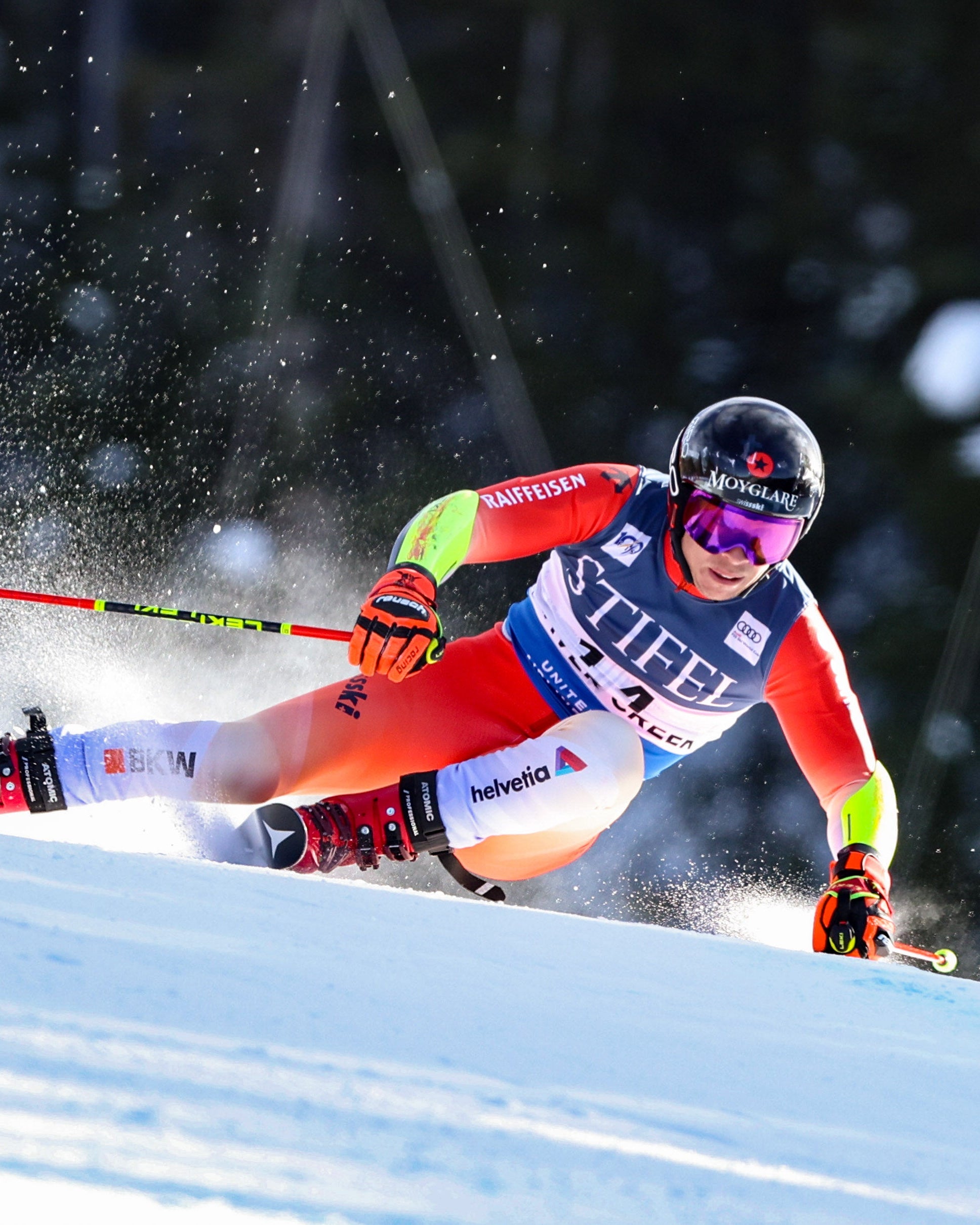 Gino Cavizel carving through fresh snow on a downhill slope, wearing a bright red, orange, and white racing suit with sponsor logos, along with a black helmet and goggles.