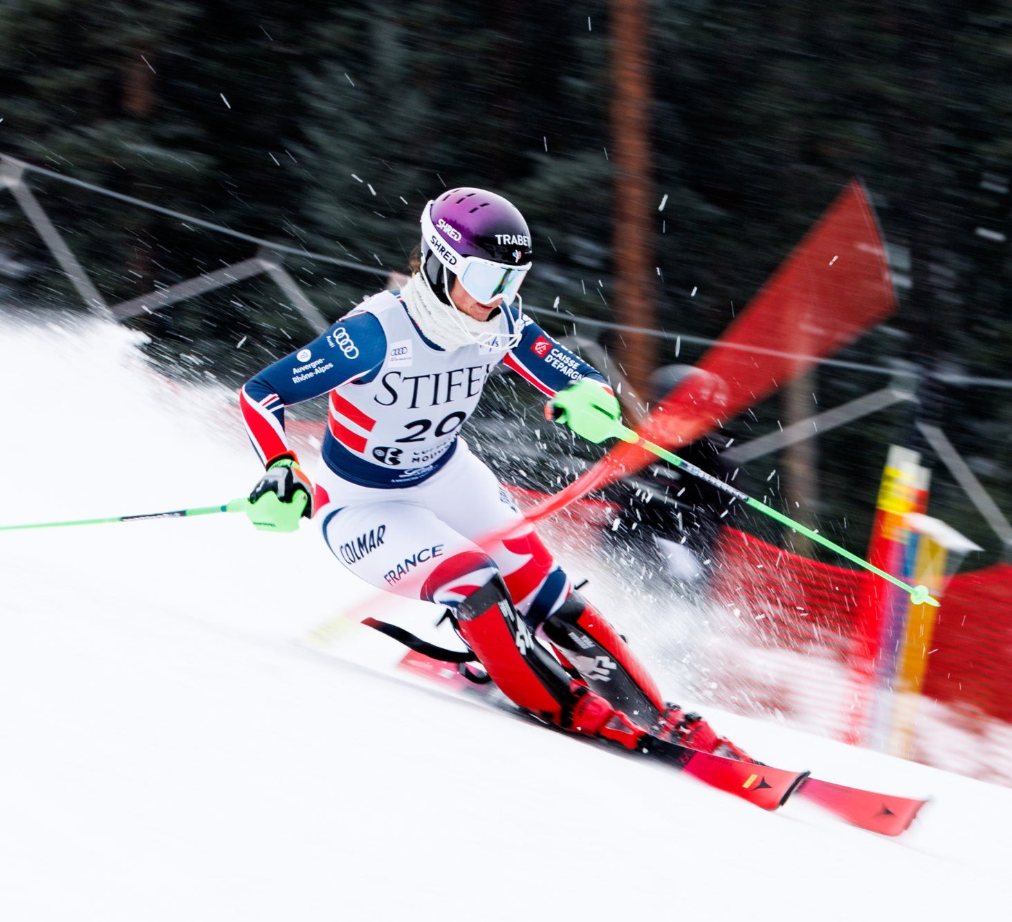 Chevrier Marion in a white, blue, and red racing suit with 'STIFFE' and '20' printed on the front, wearing a purple helmet and goggles, carving through fresh snow with red skis and green poles.