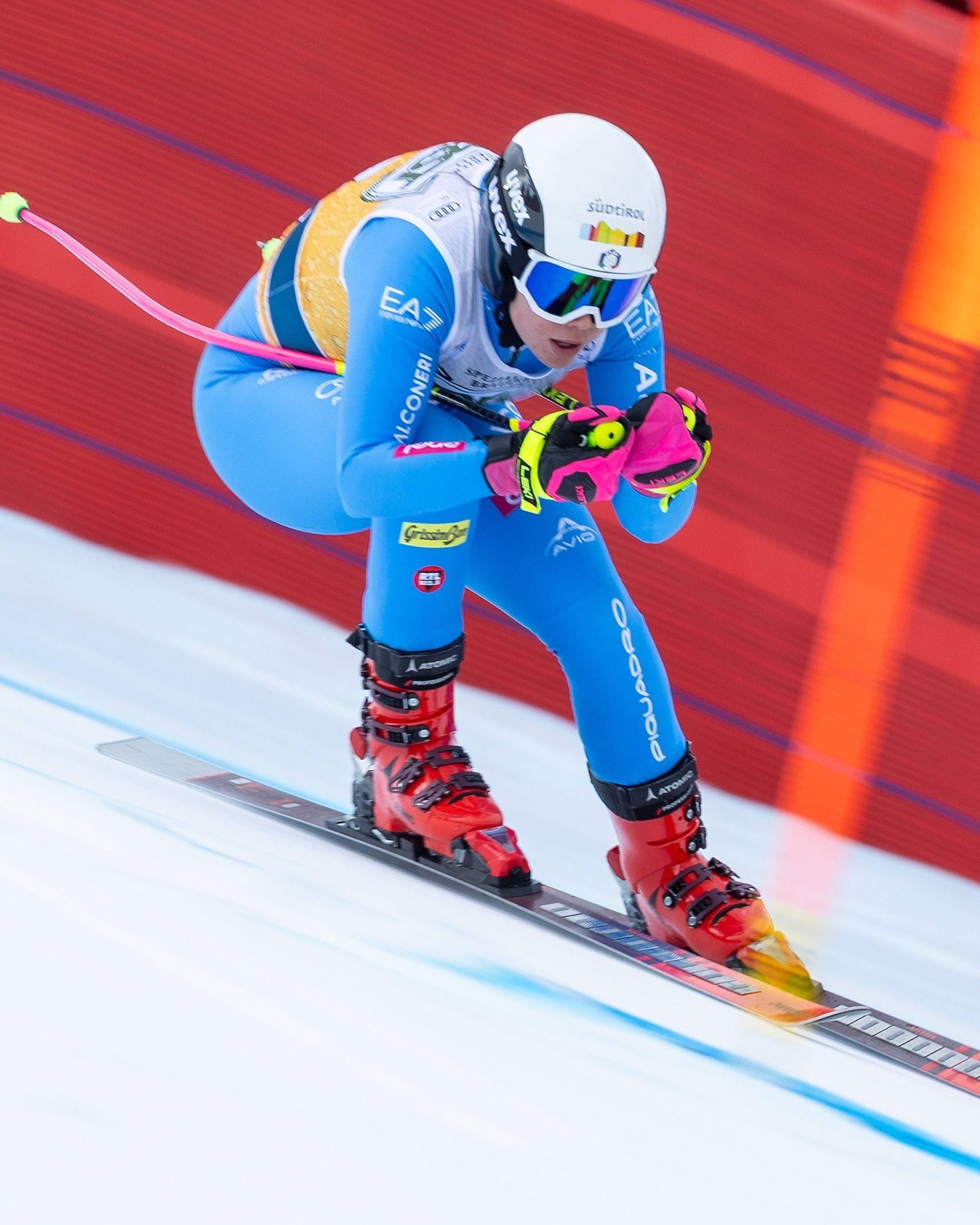 Nadia Delago in a blue and yellow suit, white helmet, and goggles leans forward on skis with pink poles, captured mid-action on a snowy track with a red and orange barrier in the background.
