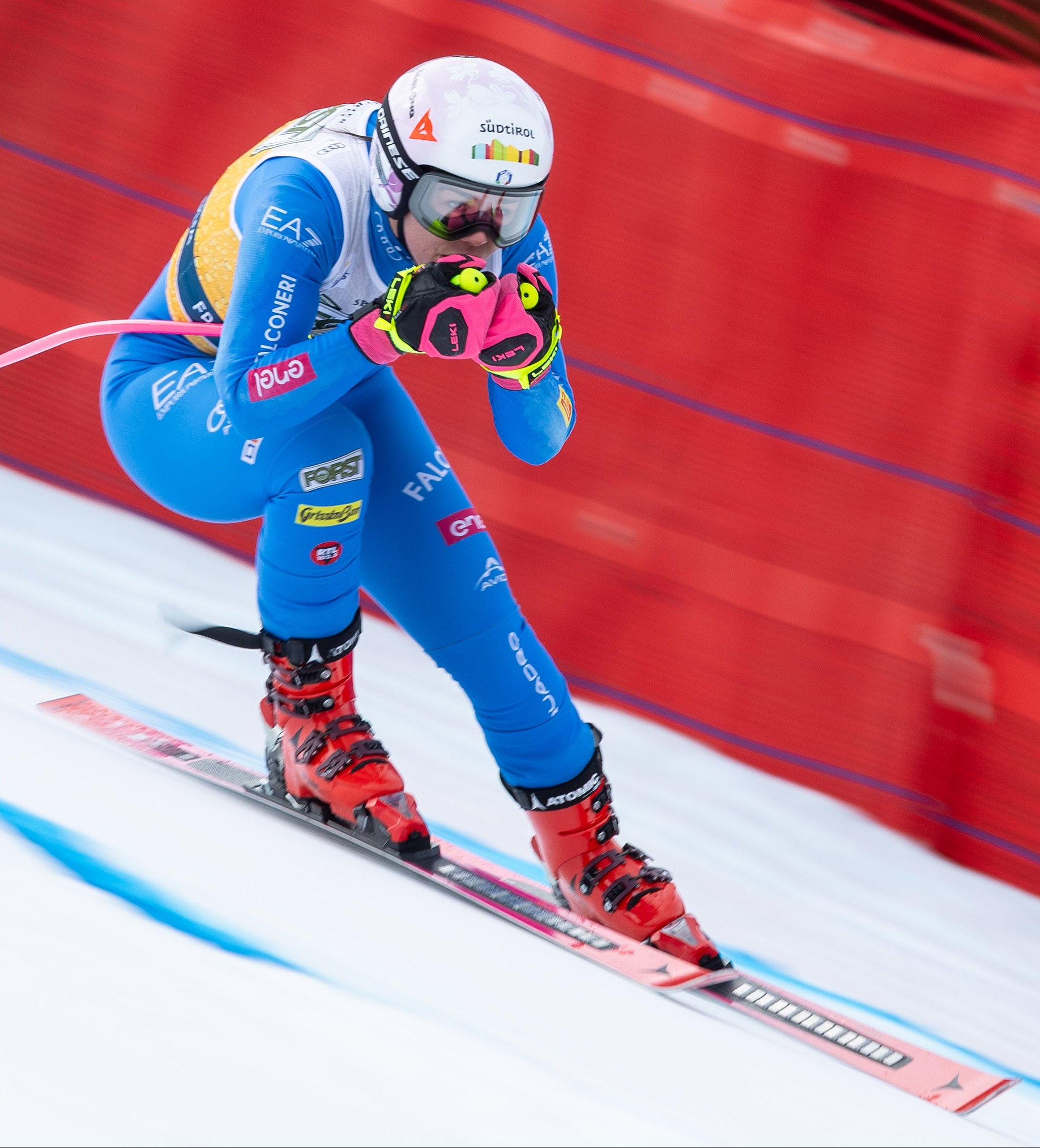 Nicol Delago in a blue and white racing suit with red boots and pink gloves carving through a snowy track, with a blurred red net in the background.