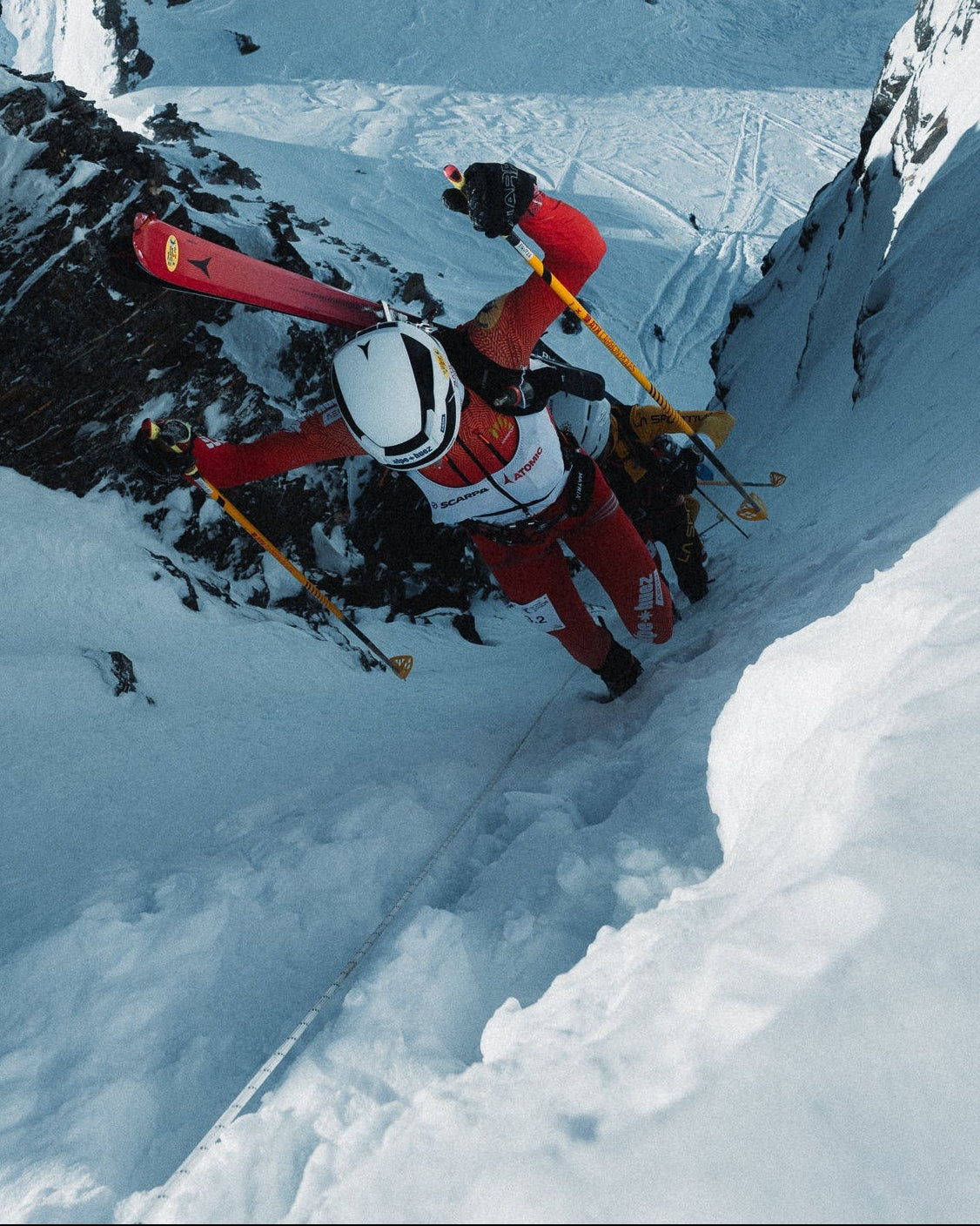 Two skiers descend a steep snowy mountain slope using ski poles and skis, with rugged snow-covered peaks in the background.