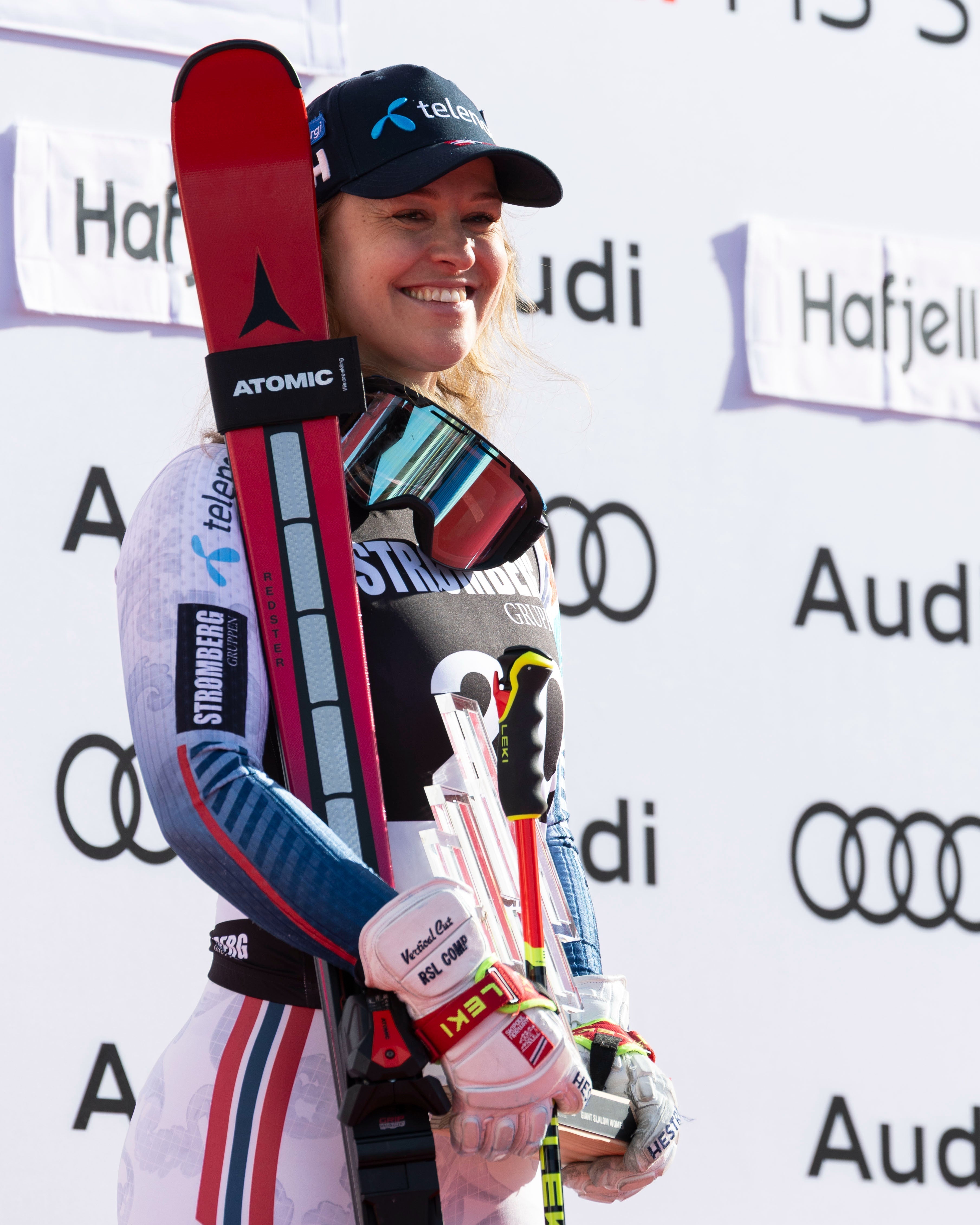Mina Holtmann stands smiling on a podium holding ski poles and a pair of skis. She wears a white, blue, and red racing suit, a black cap, and white gloves, with red and black ATOMIC skis visible.