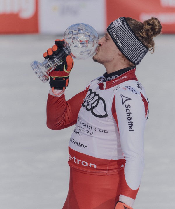 Manuel Feller in a red and white racing suit kisses the trophy after the 2024 World Cup. The skier is wearing a black and white striped beanie, orange and black gloves.