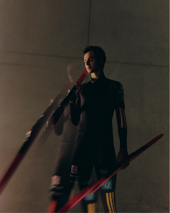 Vinzenz Geiger in a dark full-body suit holding red and black cross country skis, standing against a plain concrete wall in dim lighting.