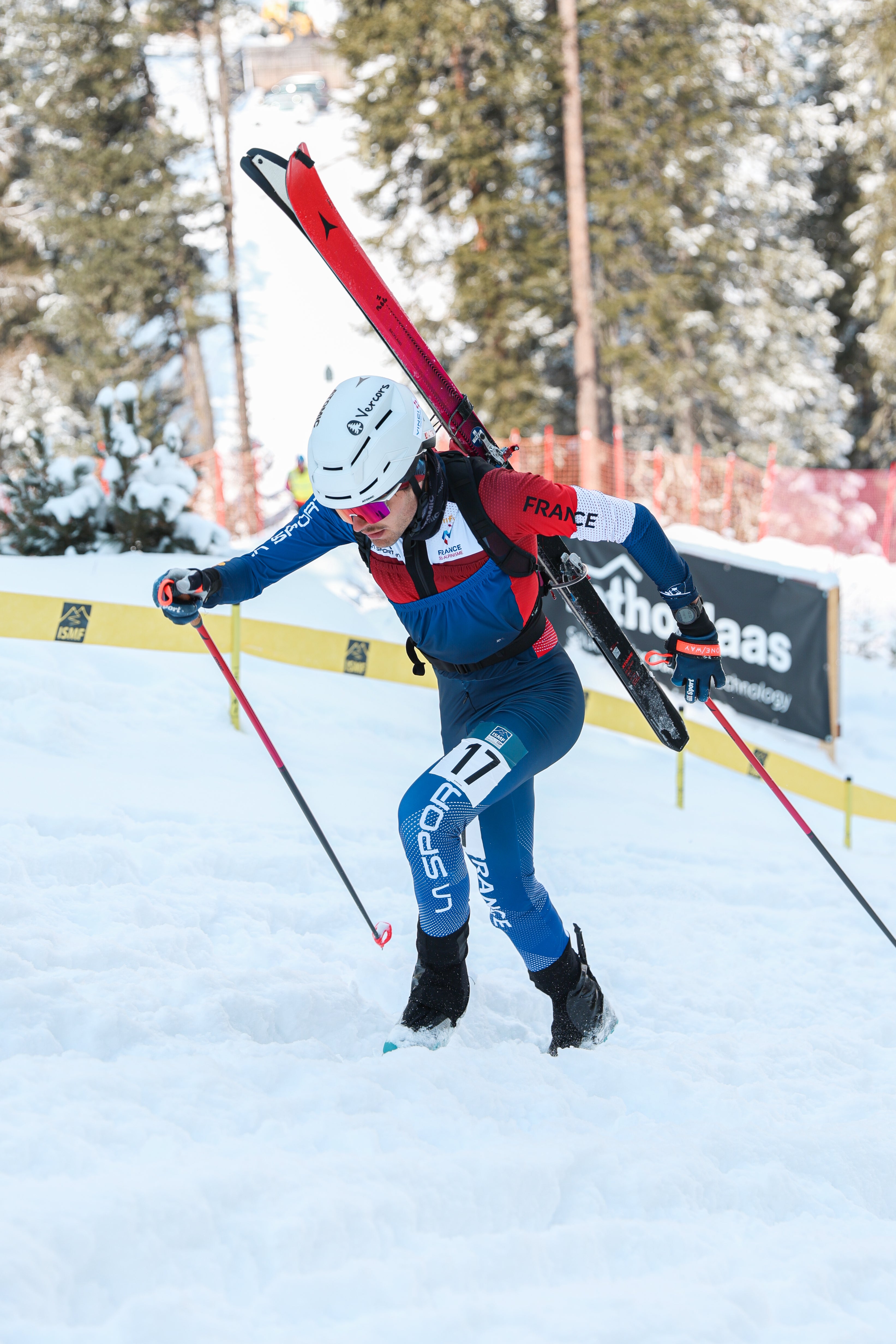 Pablo Giner in a blue, red, and white racing suit carrying red skis over their shoulders, skiing through the snow with poles in hand during a race.