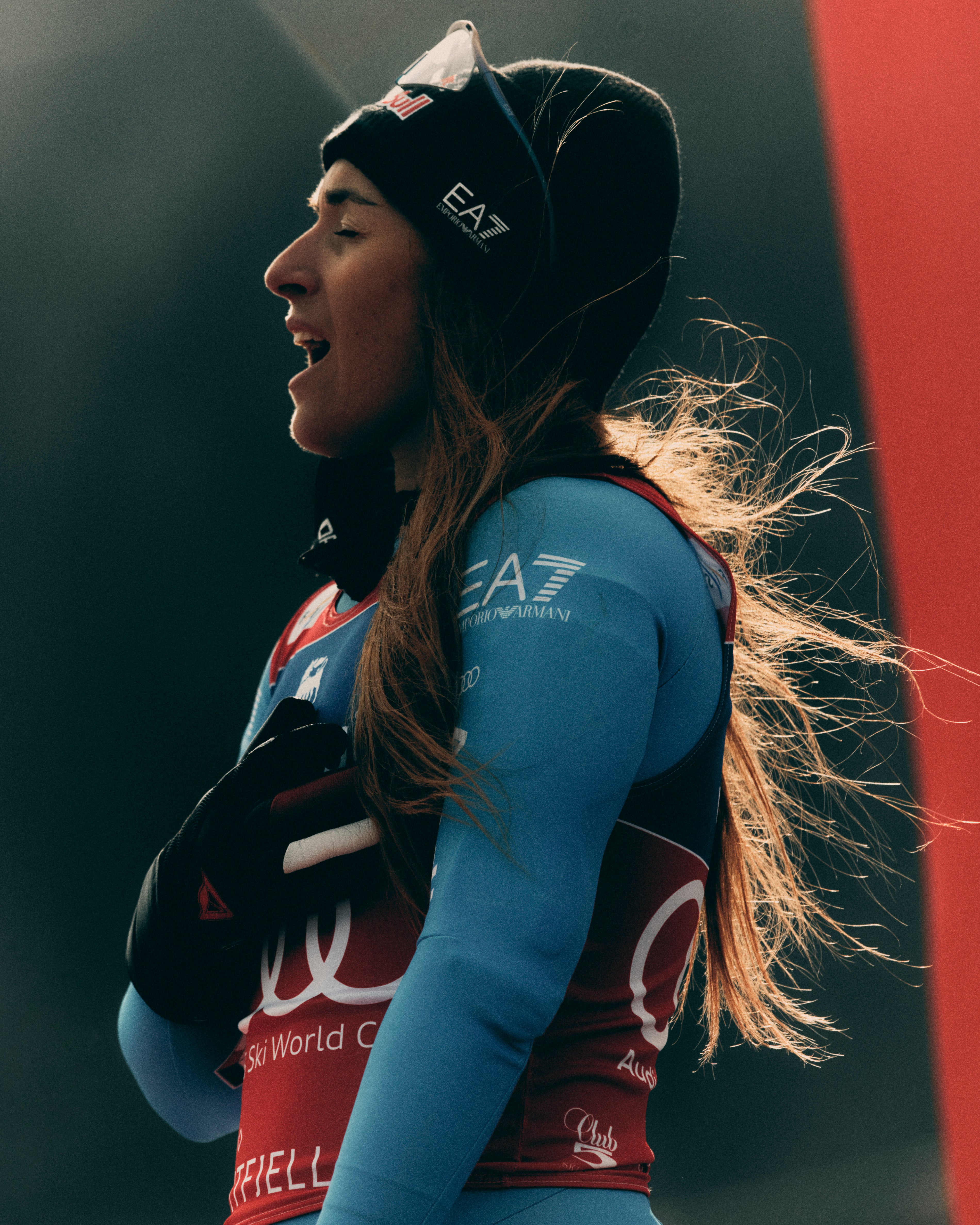 A female athlete in profile wearing a black EA7 beanie, a light blue racing suit, and a red bib with white logos, set against a dark background with a red vertical panel.