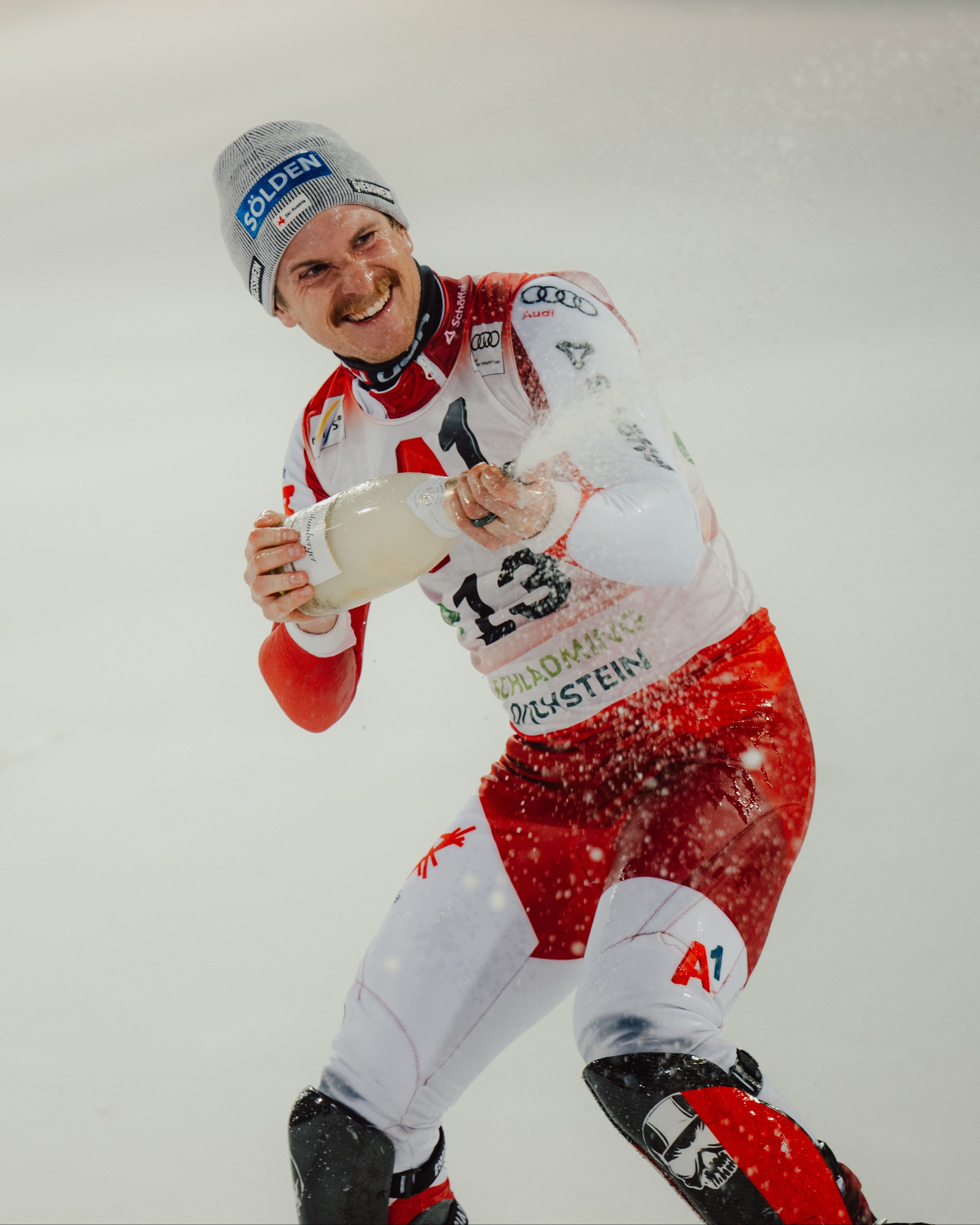 Fabio Gstrein in a red and white racing suit and gray beanie celebrates on snow while holding a bottle of champagne.