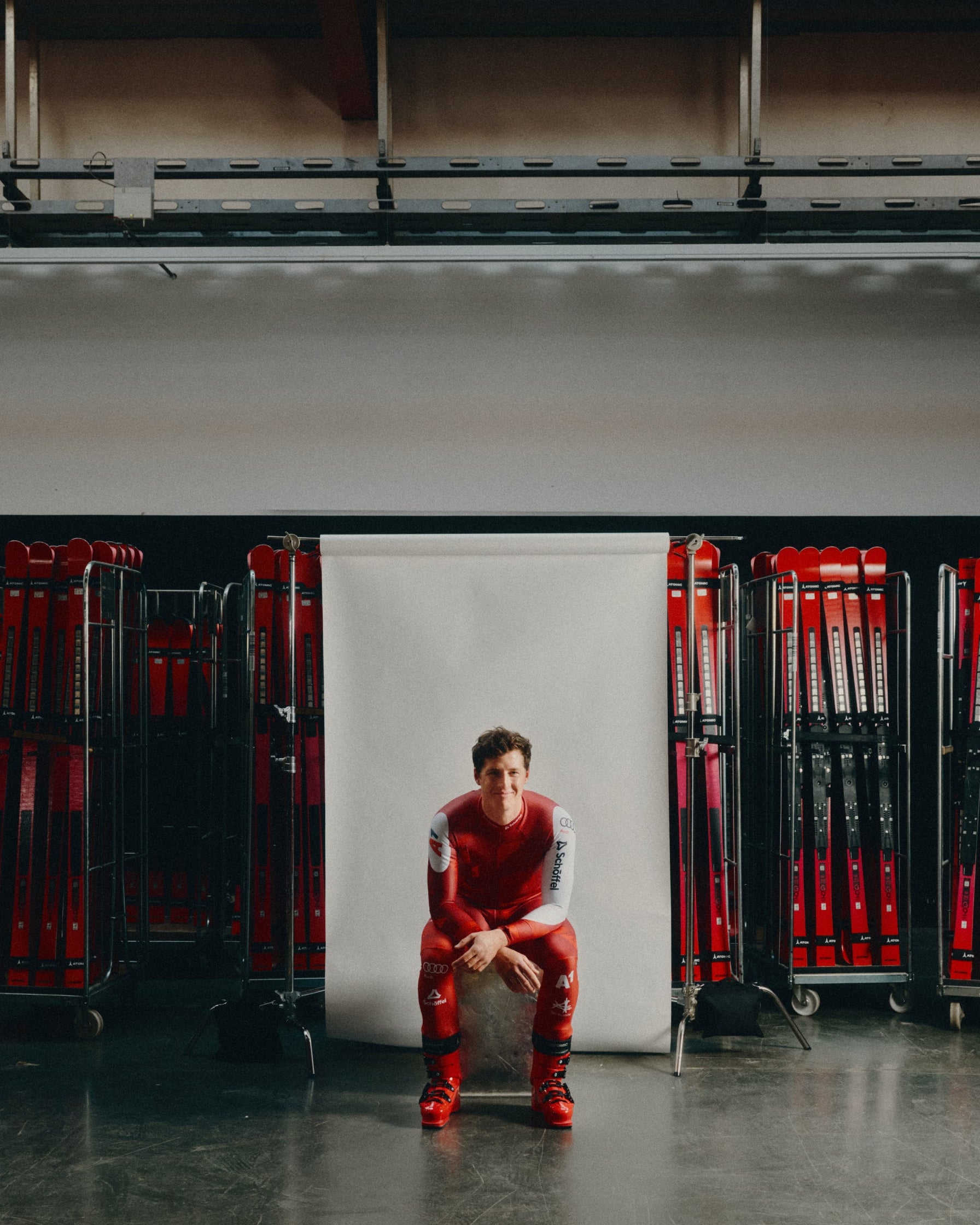 Raphael Haaser wearing a red and white racing suit sits on a chair in front of a white studio backdrop, with racks of red skis on both sides in an indoor studio setting.