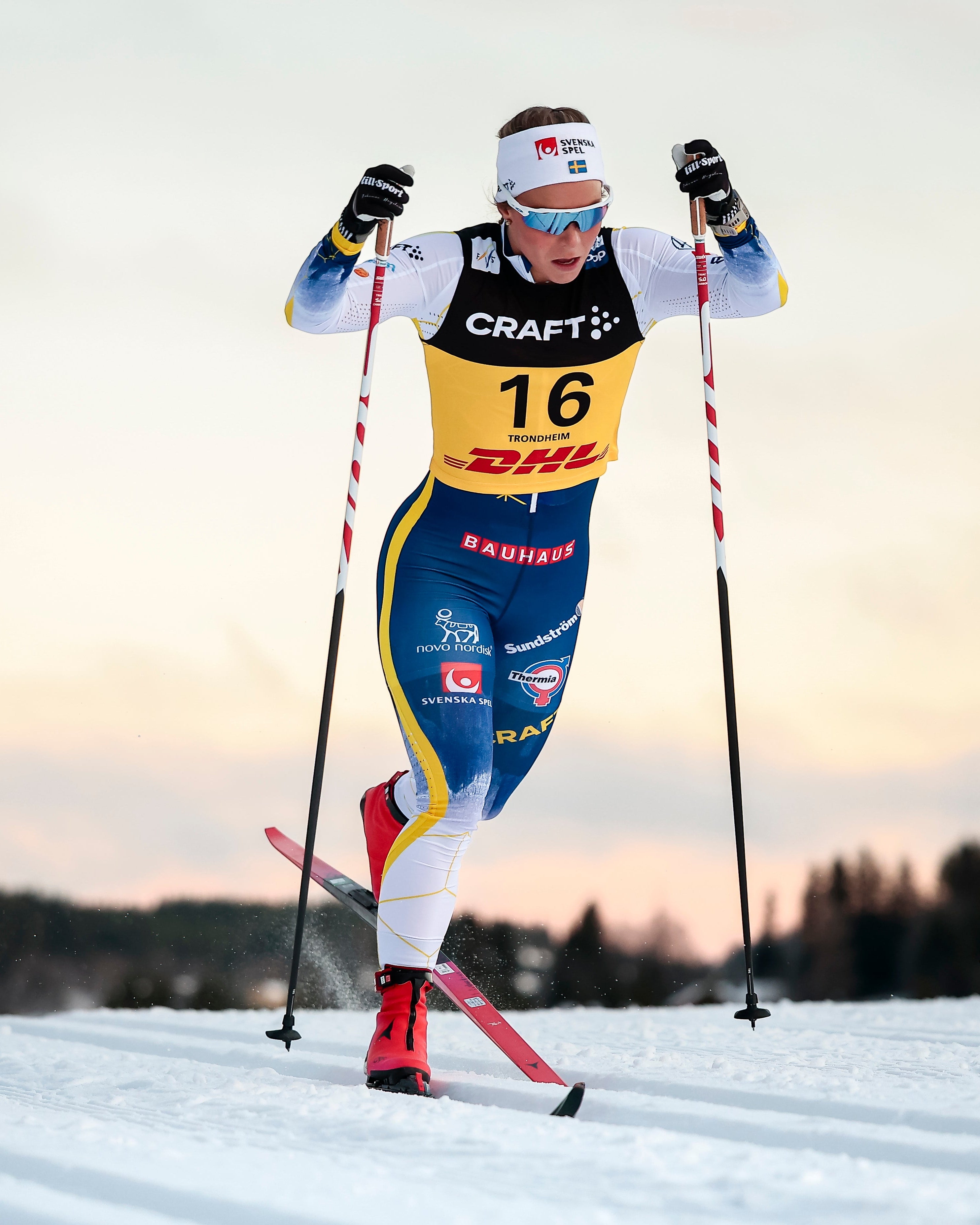 Johanna Hagstroem in a blue, yellow, and white racing suit with the number 16, skating on a snowy track during a cloudy sunset or sunrise.