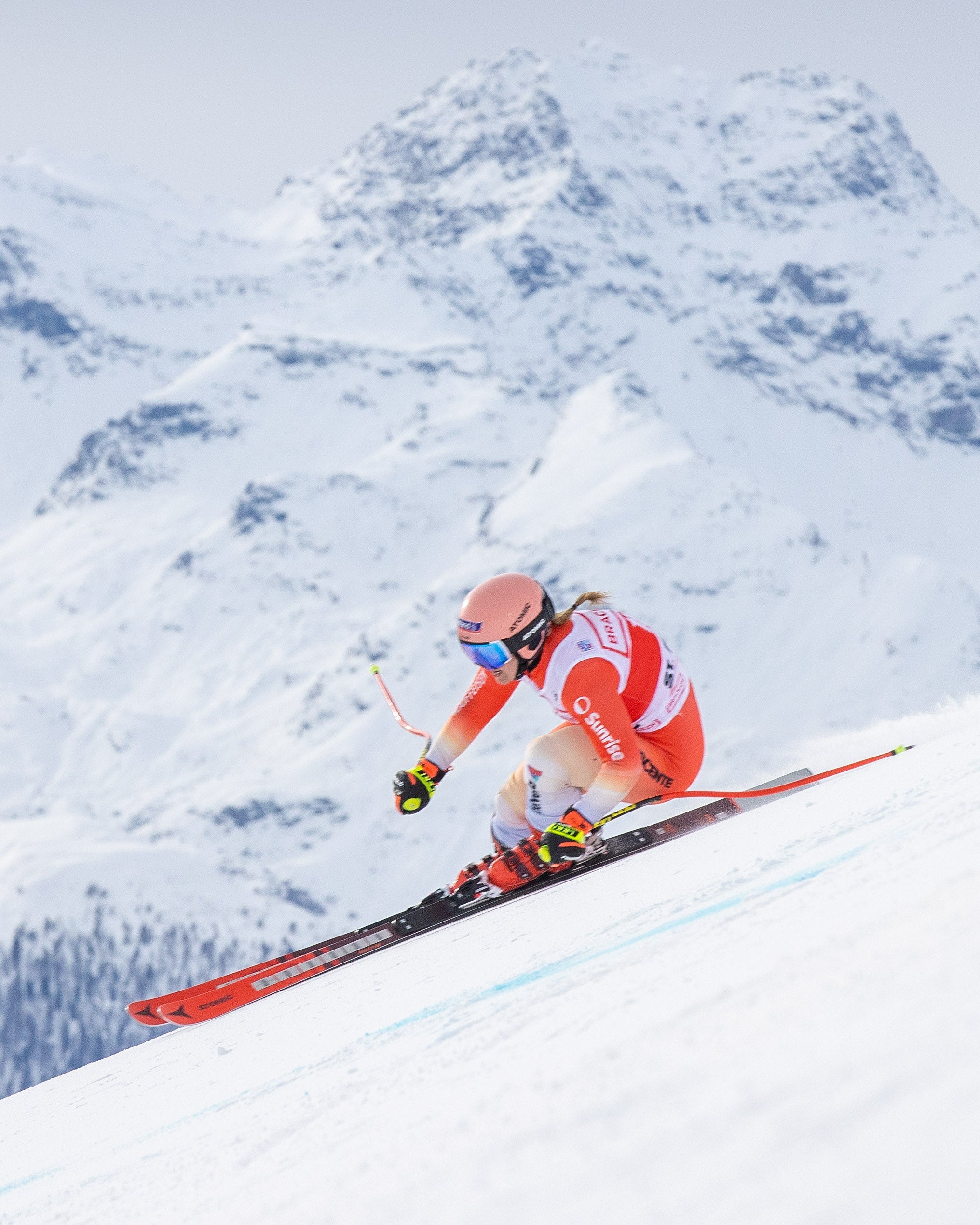 Joana Hählen in a red and white racing suit leans into a turn on a snowy mountain slope, with snow-covered peaks in the background.