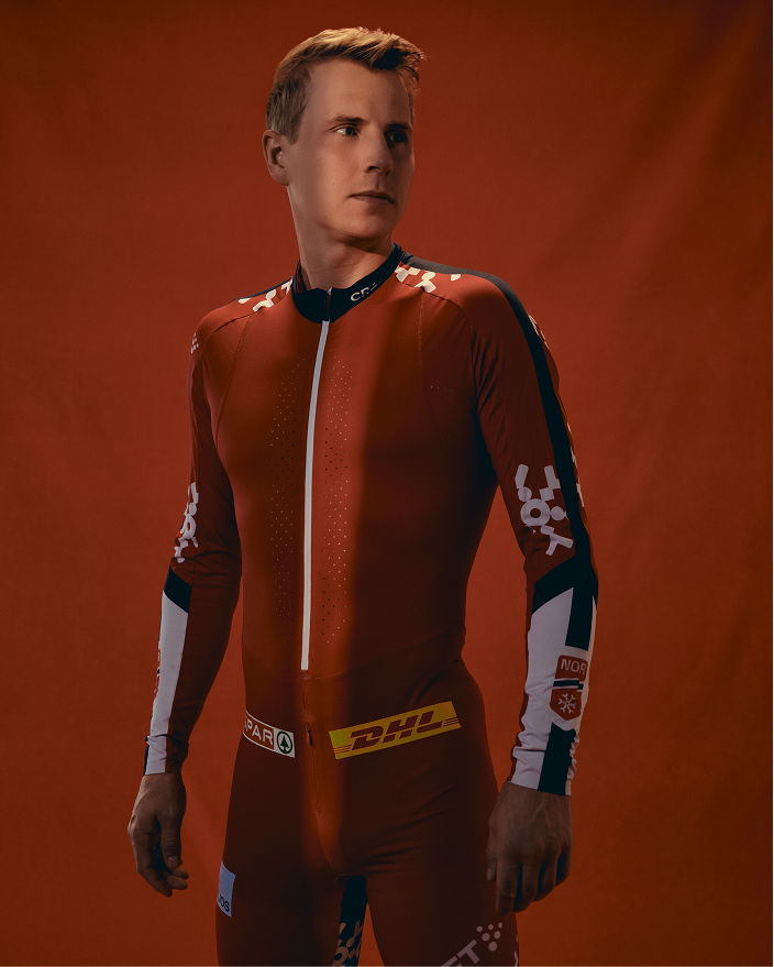 Simen Hegstad Krüger wearing a tight-fitting red racing suit with white and black sleeve panels, a white front zipper, and visible branding, posed against a warm orange-red studio backdrop.